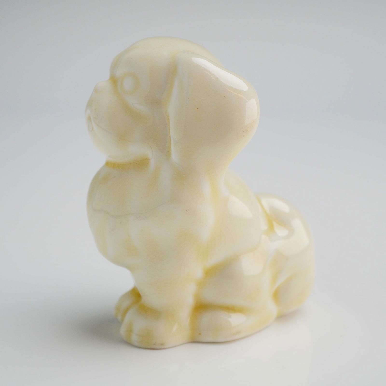Doulton England Seated Pekinese Puppy Model HN832, Rare White Colourway: A single glazed ceramic figurine manufactured by Doulton, England. The piece depicts a stylized sitting Pekinese dog. finished in a high-gloss, pale cream or buttery yellow glaze. The glaze exhibits
