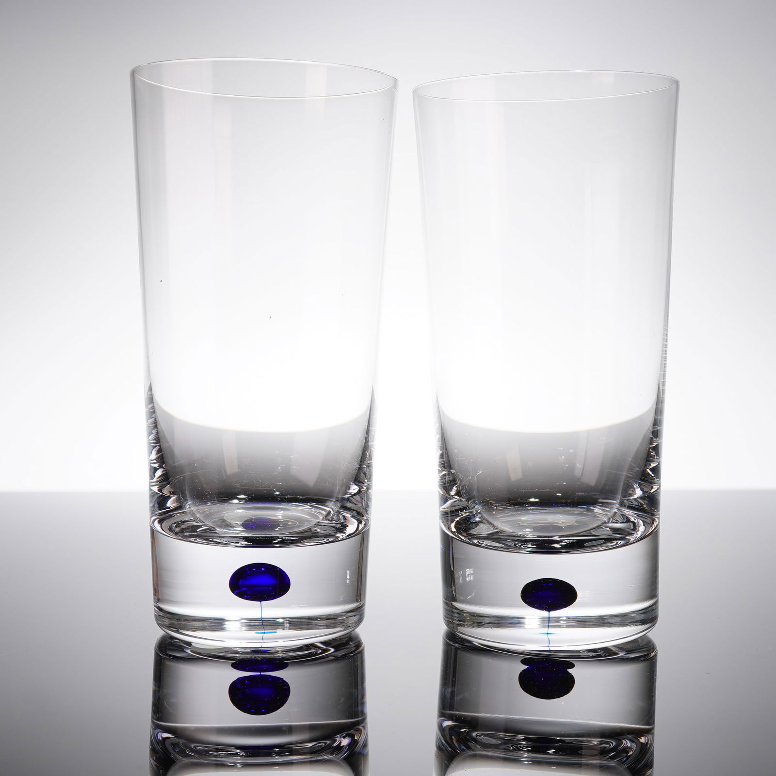 A Pair of Orrefors Intermezzo Blue Crystal Highball Tumblers: A pair of contemporary highball tumblers manufactured by Orrefors in the Intermezzo Blue patern. The glasses are crafted from clear crystal glass in a tall, cylindrical form with heavy, solid bases. T