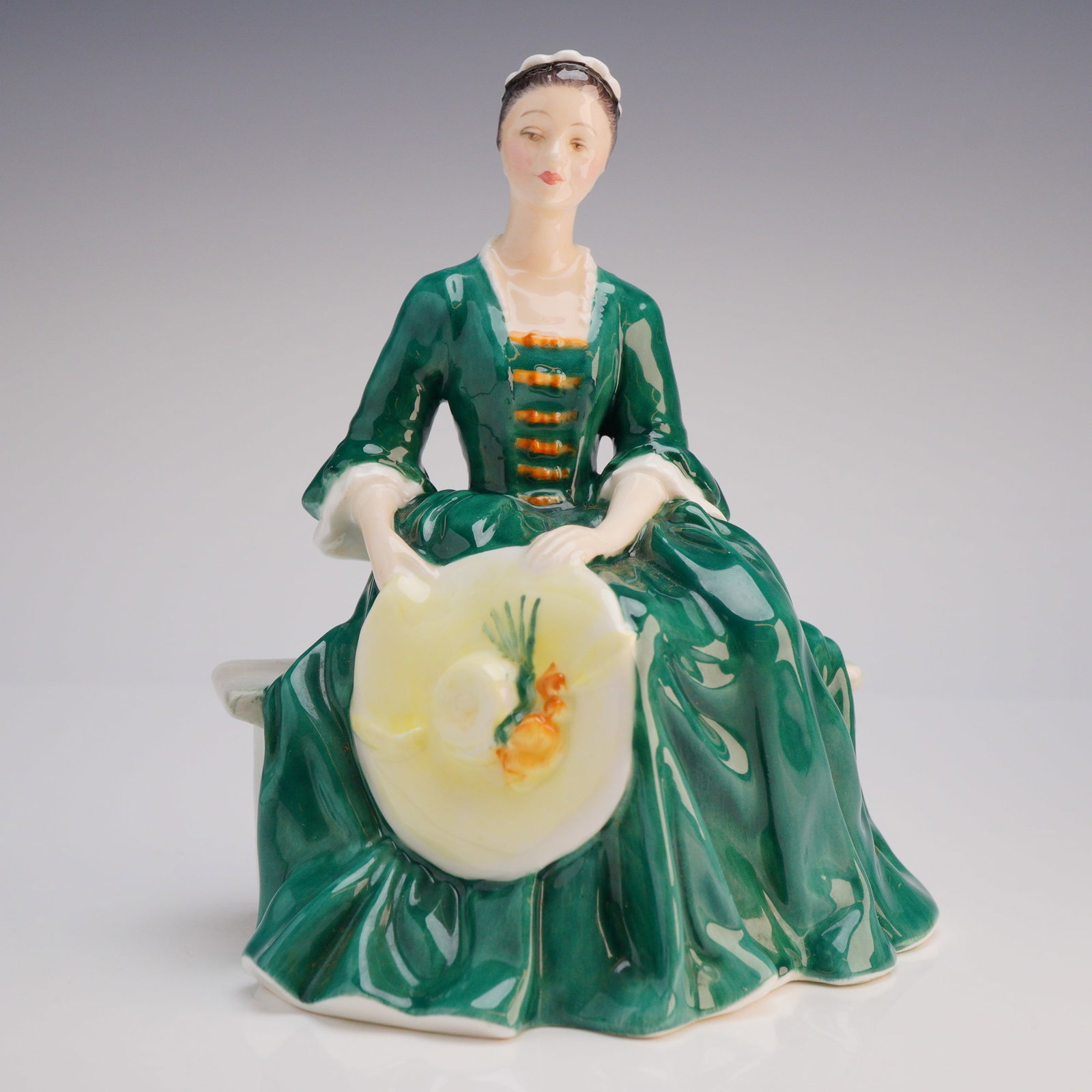 Royal Doulton Figurine A Lady from Williamsburg HN2228: The figure depicts a seated woman wearing a historically styled high-gloss green dress accented with white lace trim and orange bodice details, holding a wide-brimmed yellow hat decorated with painted