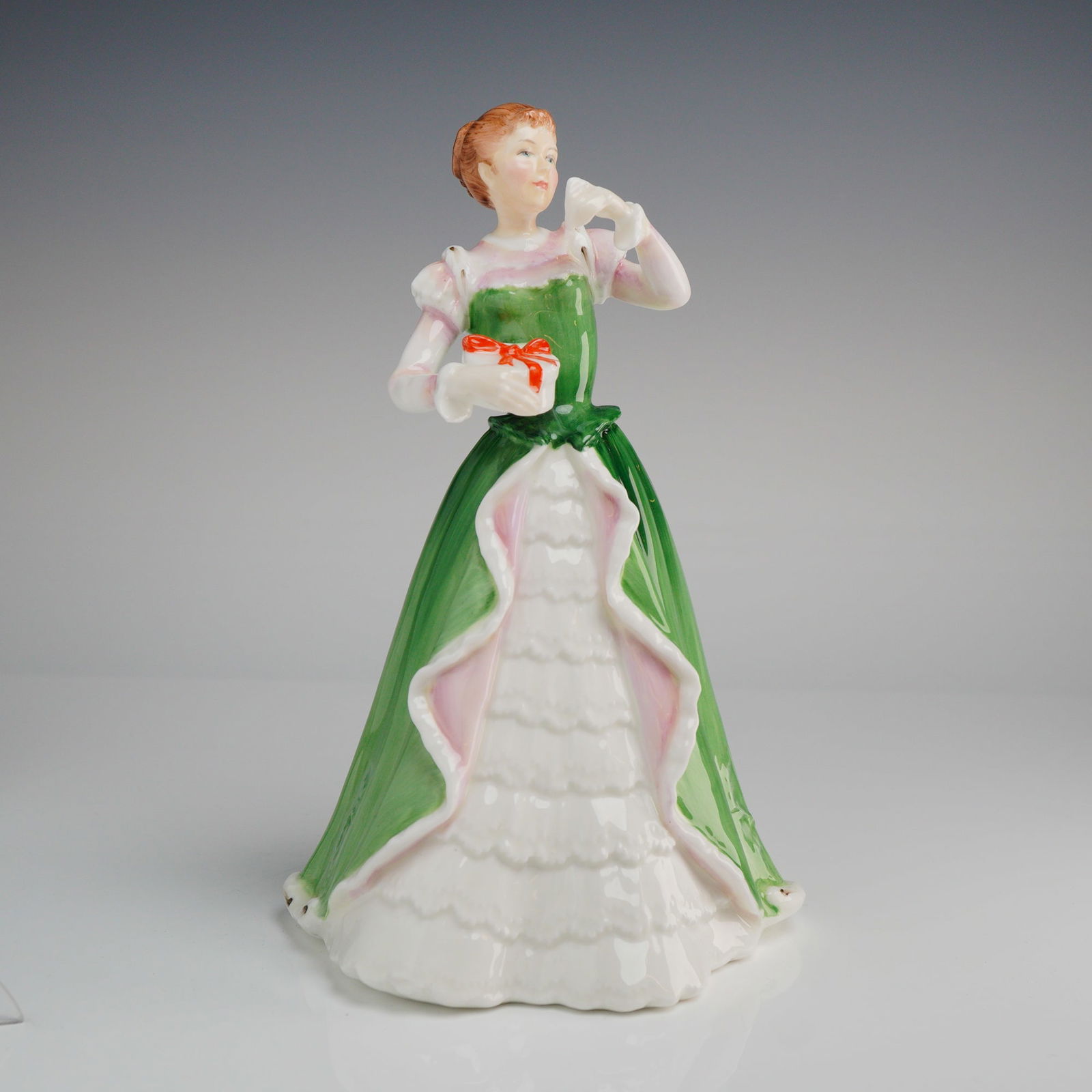 Royal Doulton Figurine, Merry Christmas HN3096: A Royal Doulton Merry Christmas HN3096 figurine, crafted from glazed bone china and hand-decorated. The figure depicts a woman in a long, flowing Victorian or Edwardian-style gown, primarily rendered