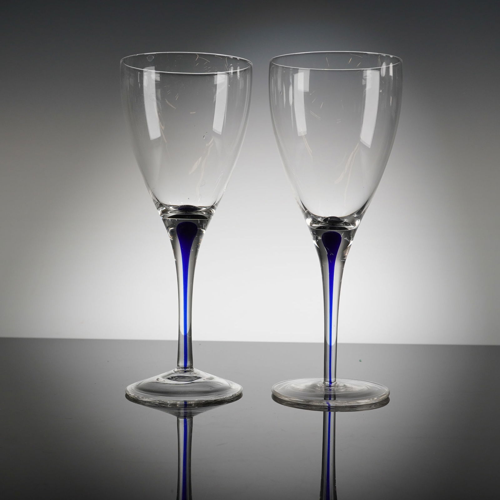 A Pair of Orrefors Intermezzo Blue Crystal Wine Glasses: A pair of Orrefors Intermezzo Blue crystal glasses. Each piece features a clear glass conical bowl set upon a tall, tapered stem and a wide circular foot. The defining characteristic of the Intermezzo