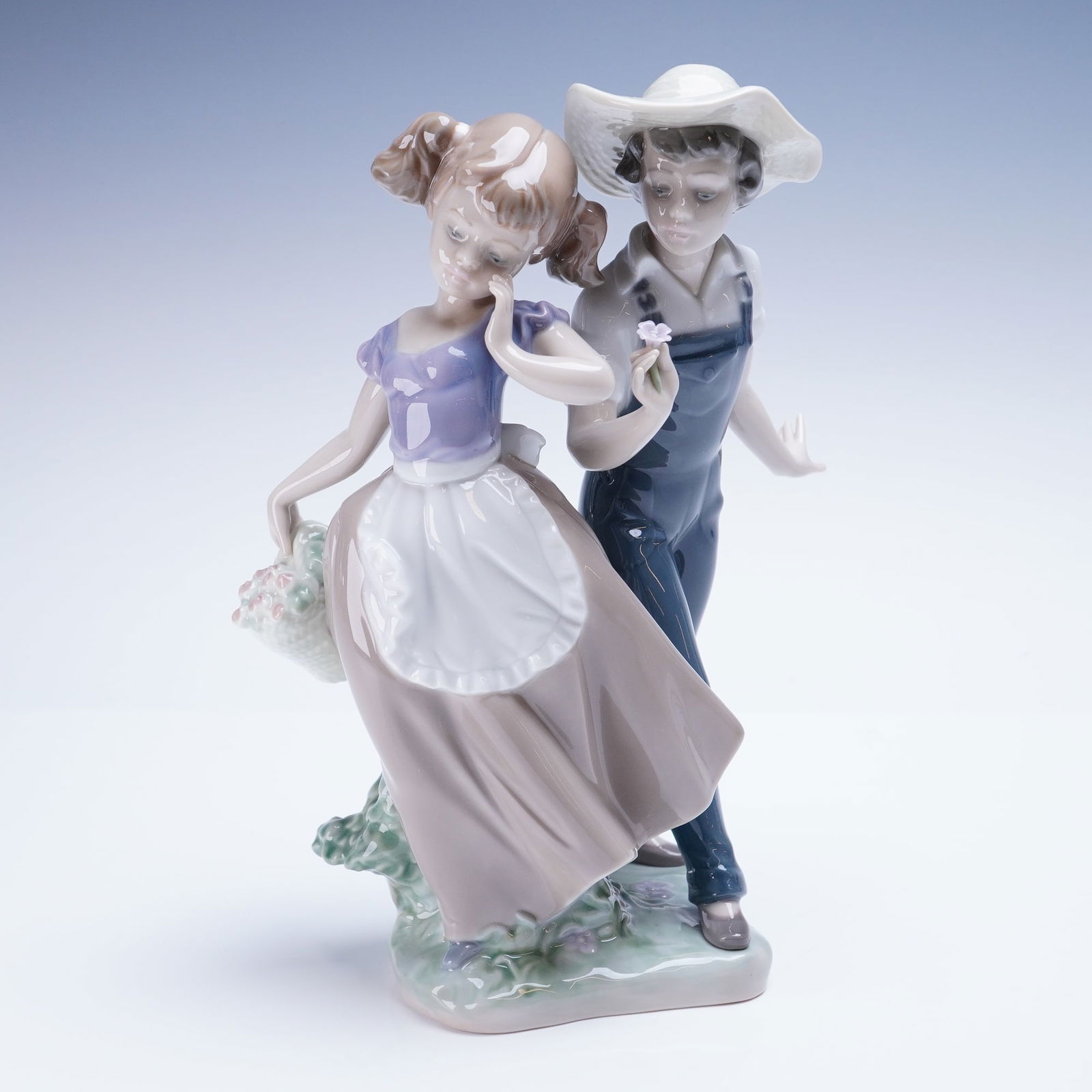 Lladro Porcelain Figurine Love in Bloom 01005292: This Lladro porcelain figurine depicts two young figures: a boy wearing blue overalls and a white hat offering a small purple flower to a girl dressed in a purple top, white apron, and taupe skirt, wh