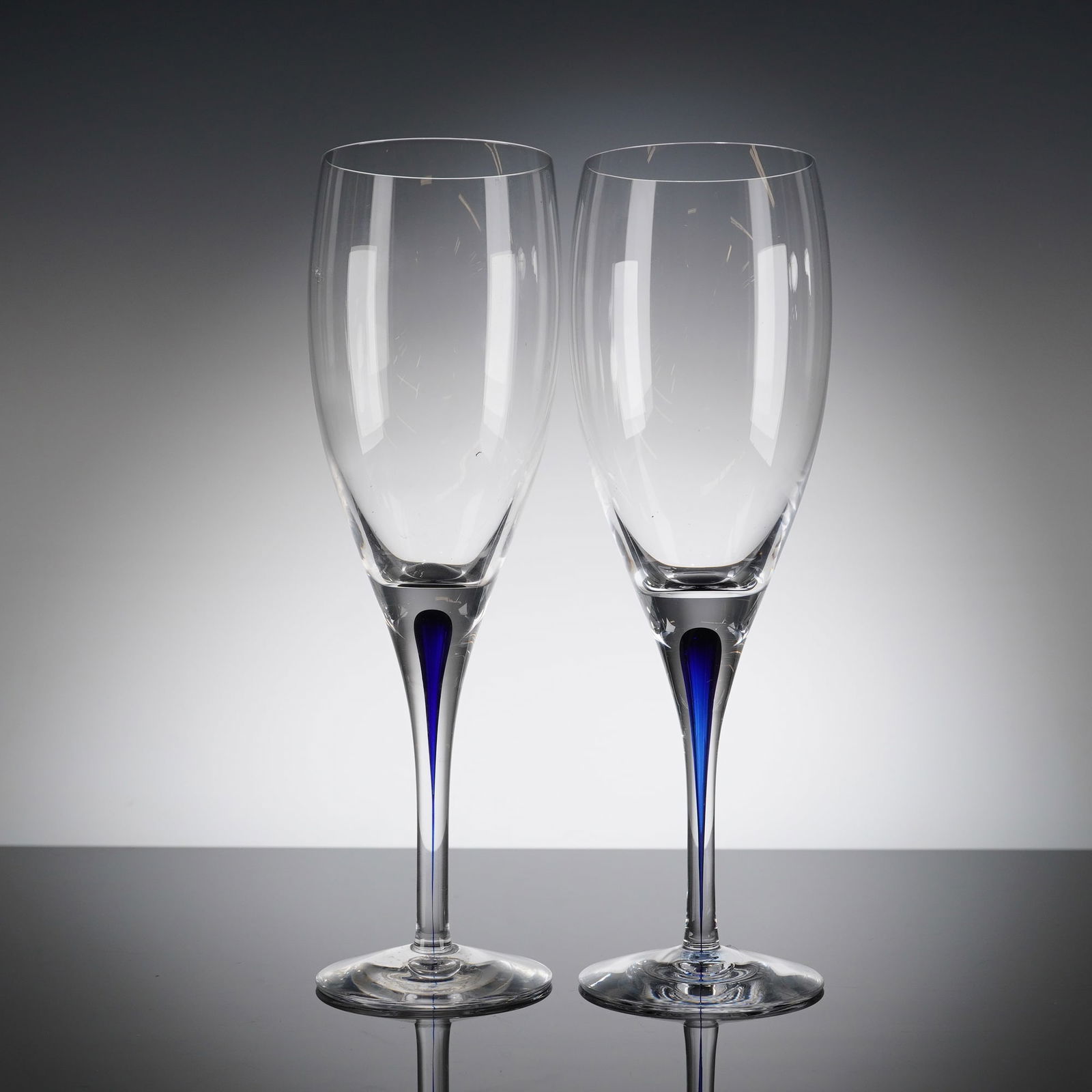 A Pair of Orrefors Intermezzo Blue Crystal Flutes: A pair of Orrefors Intermezzo Blue crystal stemware glasses. This 2pc set is executed in clear, high-quality crystal, featuring a tall, slightly tapering bowl, a slender stem, and a clear circular foo