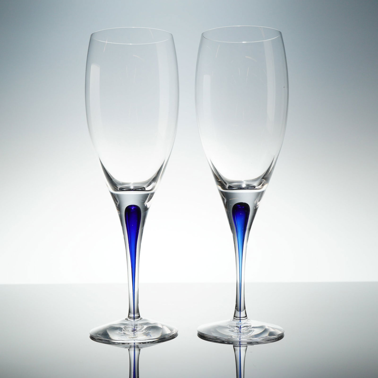 A Pair of Orrefors Intermezzo Blue Crystal Wine Glasses: A pair of Orrefors Intermezzo Blue crystal wine glasses. The 2pc set is constructed from clear, mouth-blown glass, featuring a tapering bowl set upon a slender stem. The distinctive design incorporate