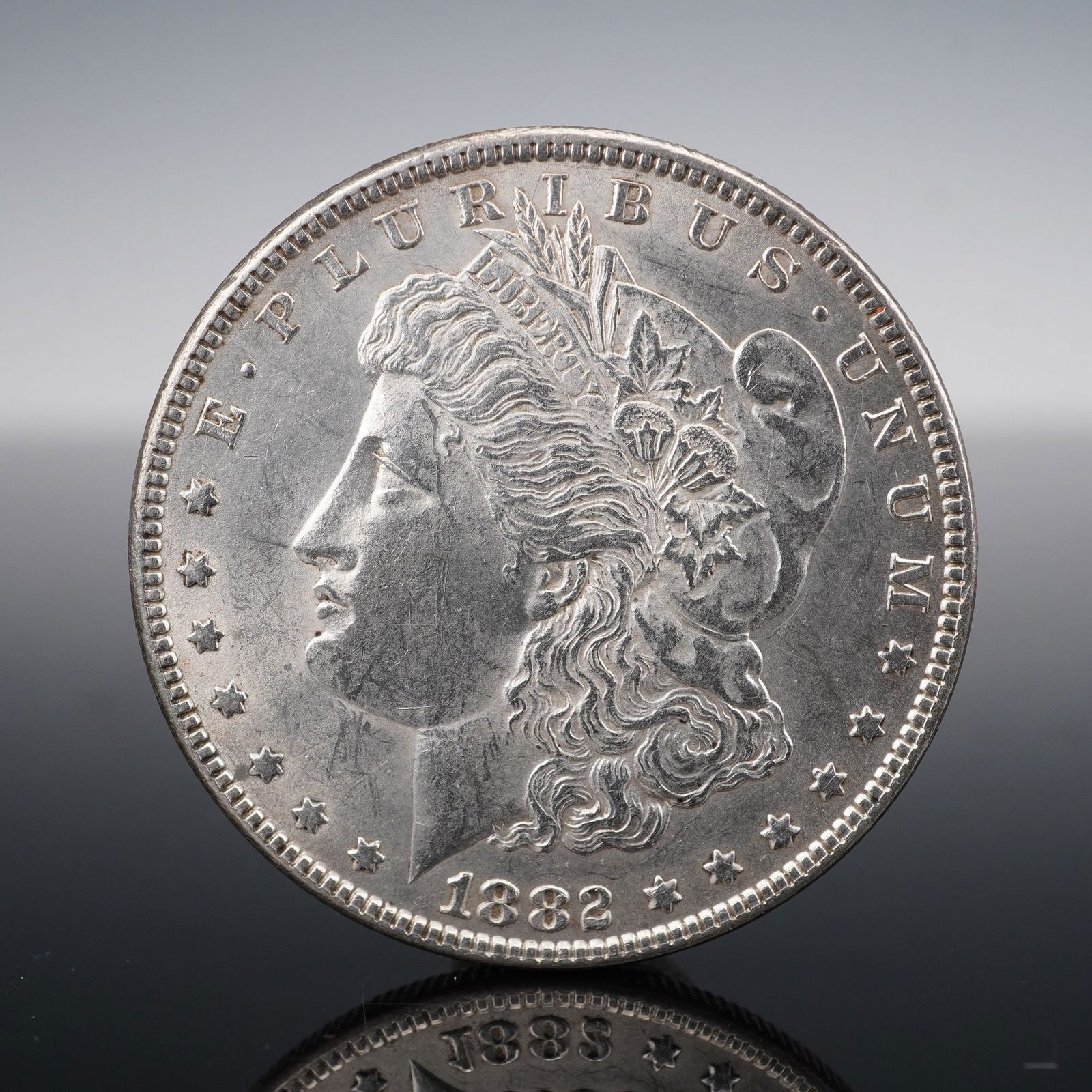 1882 Morgan Silver Dollar: An 1882 Morgan Silver Dollar, struck in ninety percent silver, designed by George T. Morgan. The obverse features a left-facing portrait of Lady Liberty adorned with a Phrygian cap, cotton blossoms, a