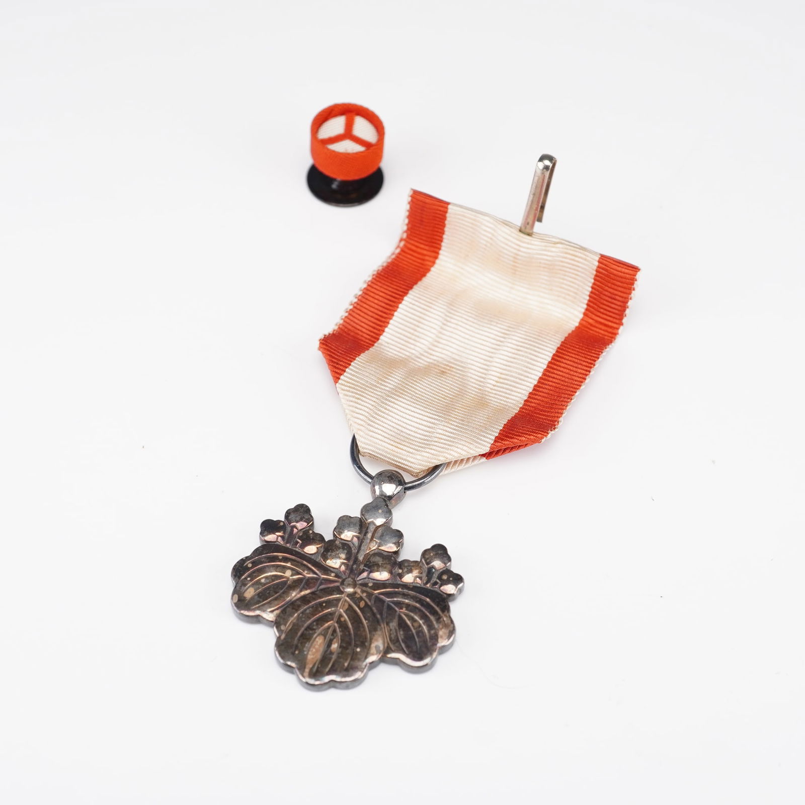Japanese Order of the Sacred Treasure Medal in Presentation Case: Medal presented in its original fitted case lined with purple silk. The medal features a silvered chrysanthemum crest suspended from a white and orange ribbon, accompanied by a matching lapel rosette.