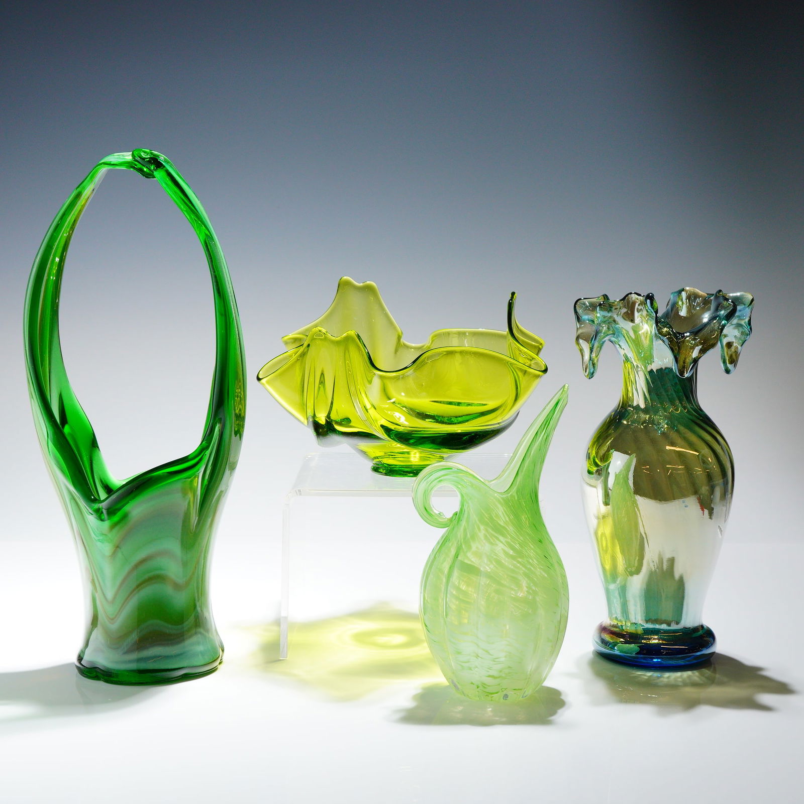 4pc Mid-Century Style Green Art Glass Vessels: A collection of four hand-blown art glass vessels dominated by various shades of green. The group includes four distinct pieces. A tall handled basket featuring swirling opaque and semi-opaque emerald