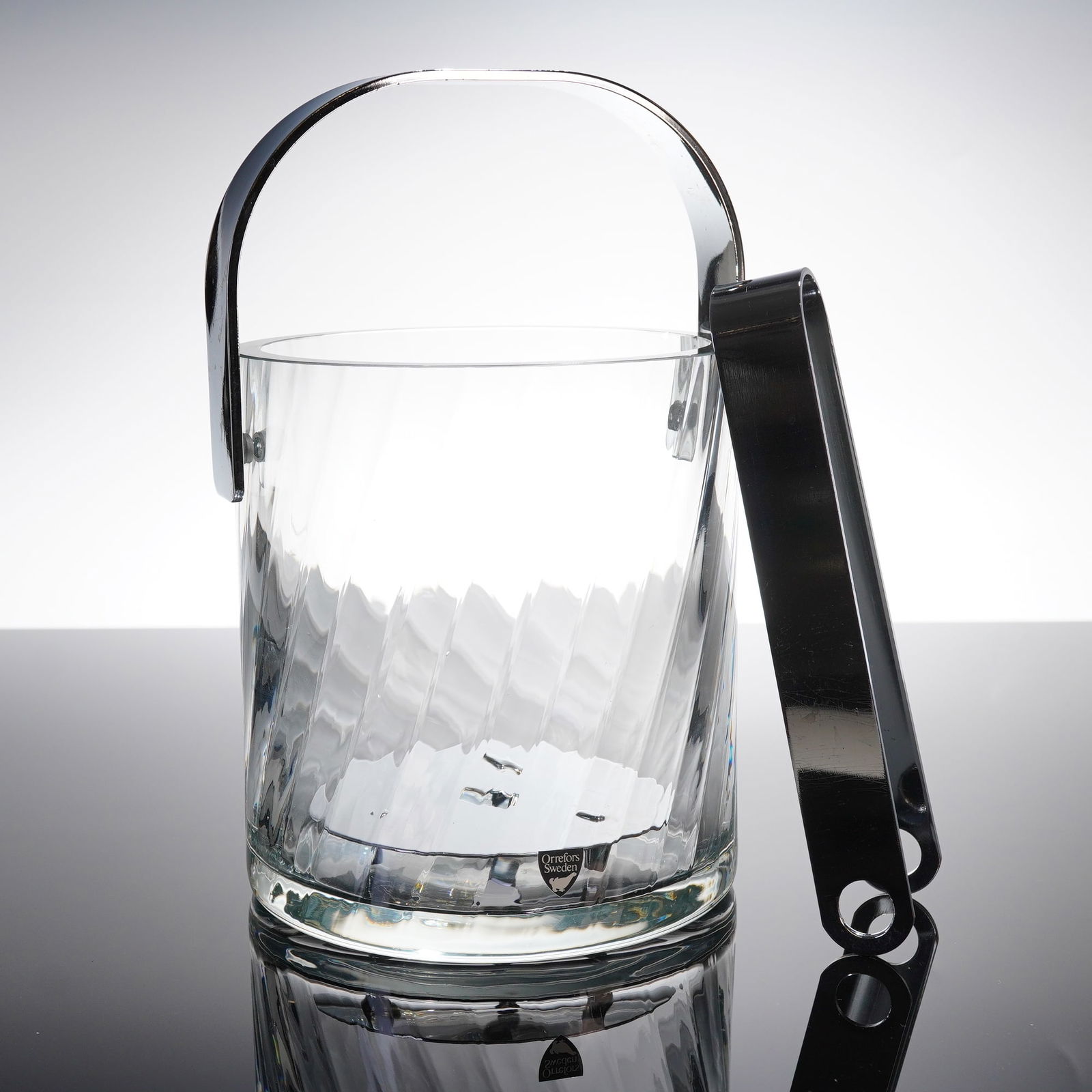 Orrefors Sweden Helena Crystal Three-Piece Ice Bucket Set: A three-piece modern serving set by Orrefors, Sweden, identified as the Helena patern. The set comprises a heavy clear crystal ice bucket, a pair of ice tongs, and a circular polished metal insert/dra