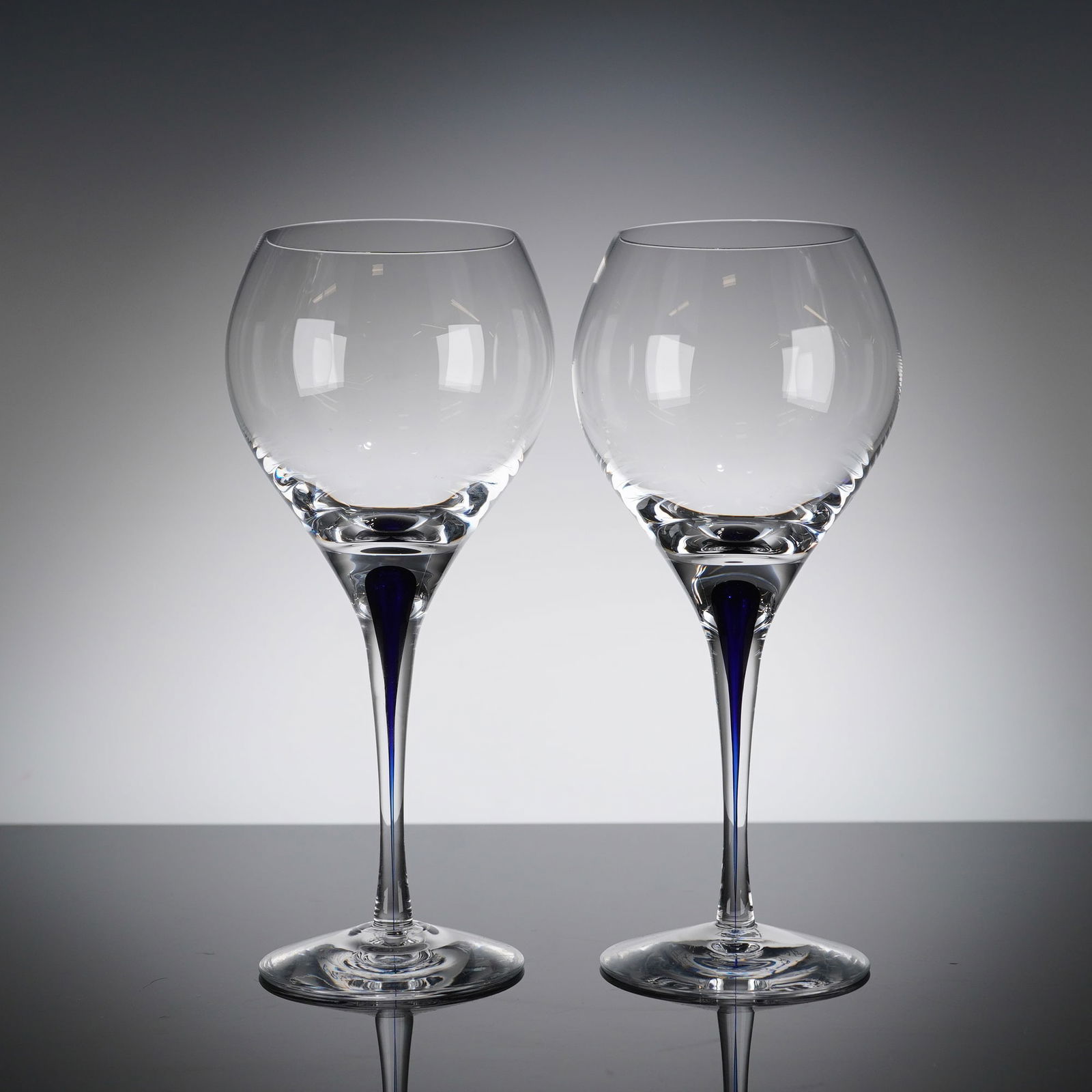 A Pair of Orrefors Intermezzo Blue Crystal Wine Glasses: A Pair of crystal wine glasses executed in the Intermezzo Blue patern by the Swedish manufacturer Orrefors. These Scandinavian modern glasses feature a large, rounded bowl set atop a clear, tall, tape