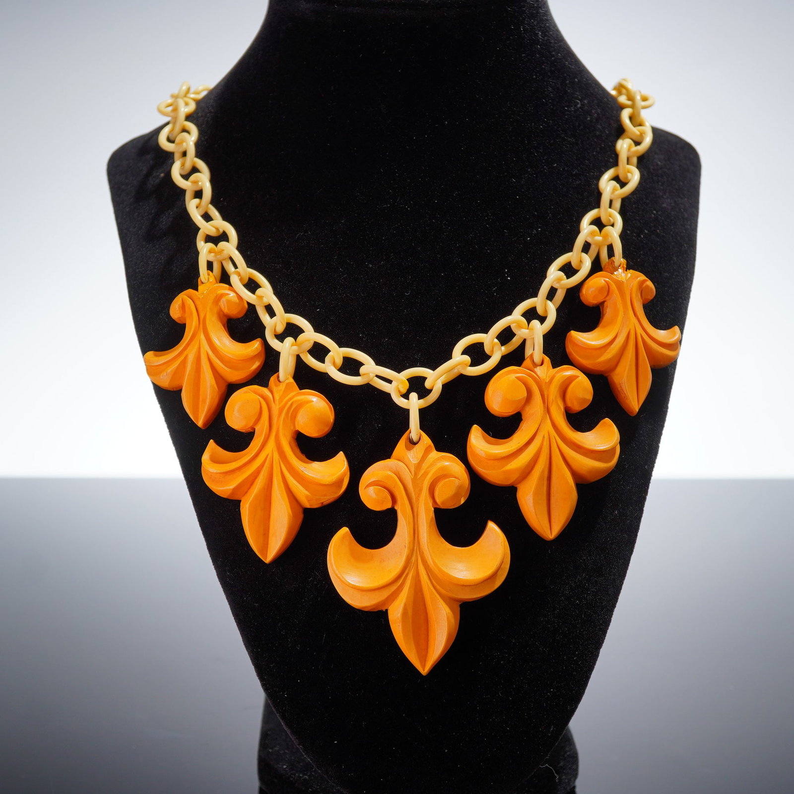 Bakelite Fleur de Lis Charm Necklace: A vintage necklace featuring a cream-colored celluloid and metal link chain. Suspended from the chain are five large, carved, light brown Bakelite charms, each deeply carved in a dramatic fleur-de-lis