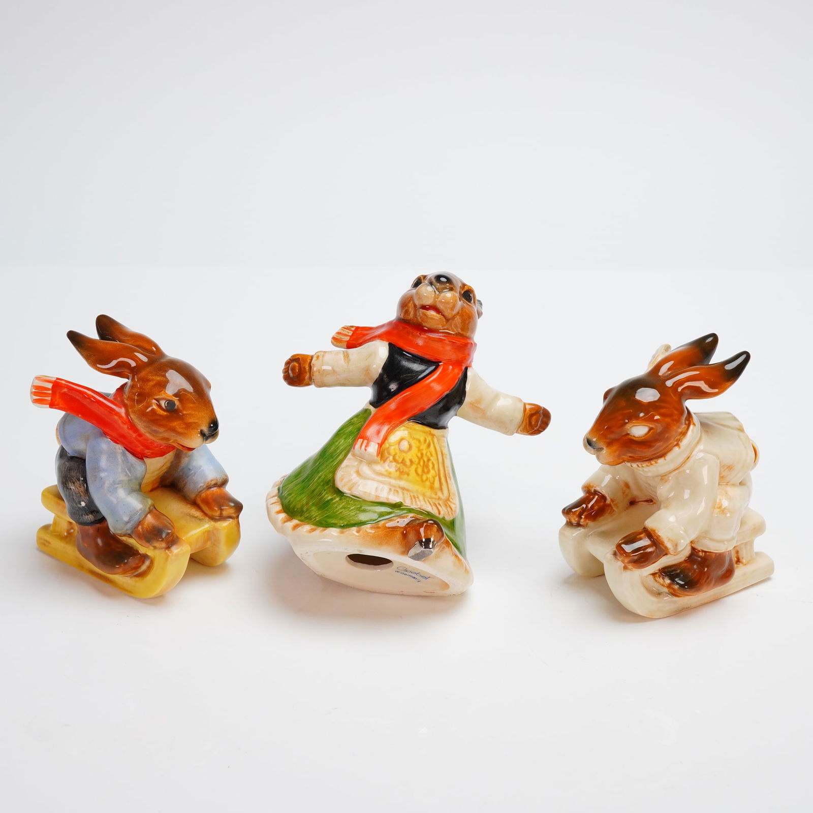 3pc Goebel Bavarian Bunnies Figurines: This trio depicts anthropomorphic Goebel Bunnies figurines engaged in winter activities. The collection includes two rabbits seated on sleds (one wearing a blue jacket, the other a cream coat) and a c