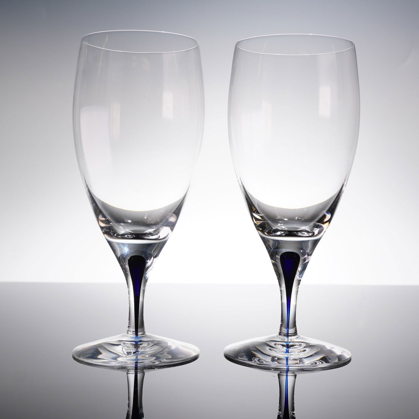 A Pair of Orrefors Intermezzo Blue Crystal Iced Tea Glasses: A matched pair of Orrefors Intermezzo Blue stemware, identified as Iced Tea Glasses. The items are executed in clear crystal or high-quality glass, featuring a tall, slightly tapered bowl supported