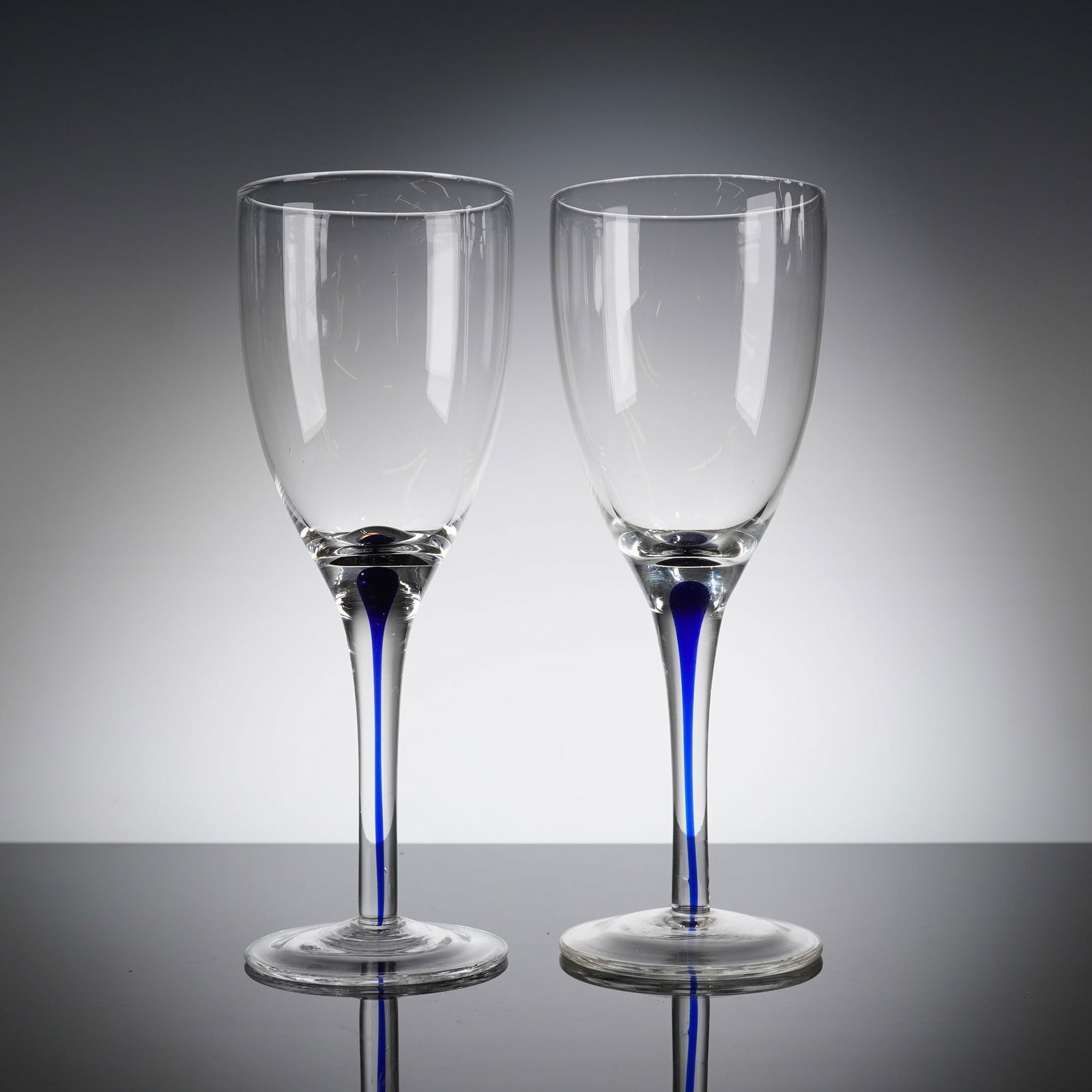 A Pair of Orrefors Intermezzo Blue Crystal Wine Glasses: A pair of Orrefors Intermezzo Blue crystal glasses. These contemporary drinkware items are mouth blown from clear, glass, featuring a tapering bowl, slender stem, and circular foot. The defining