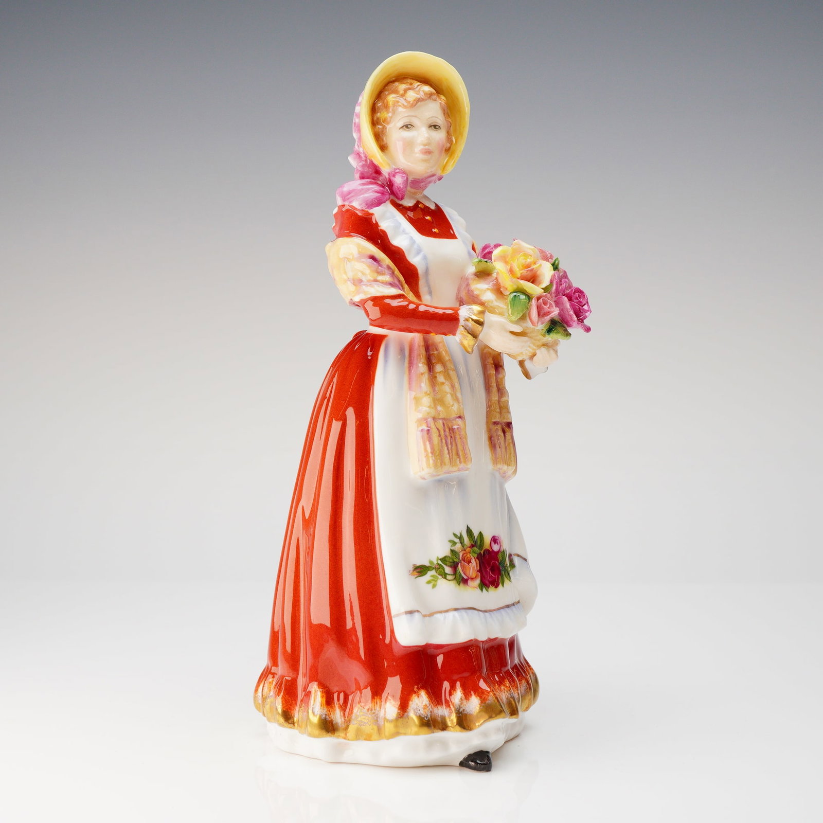 Royal Doulton Bone China Figurine, Old Country Rose HN3692: A hand-painted figure depicting a woman in a flowing red dress, complemented by a yellow bonnet with a pink ribbon and a coordinating yellow-and-pink shawl. She wears a white apron decorated with the