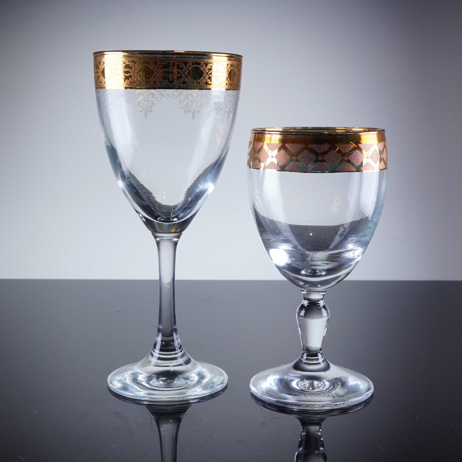 2pc Gold Rim Crystal Glassware: A group of stemware drinking glasses, comprising one water goblet and one wine glass. Both pieces are executed in clear crystal and feature extensive gold-encrusted decoration applied around the rim.