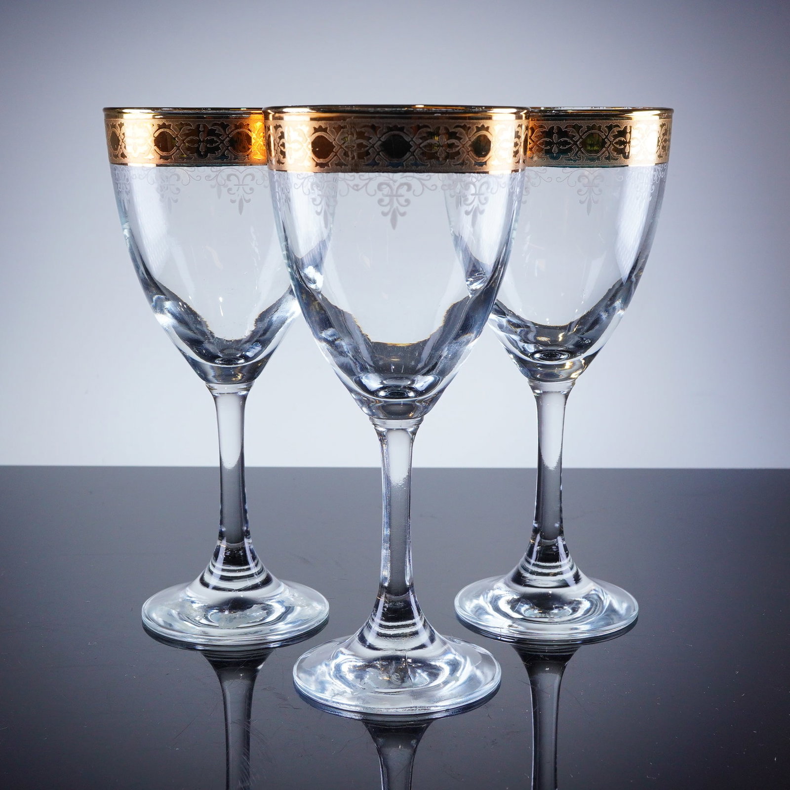 3pc Gold Rim Crystal Wine Glasses: A trio of clear crystal wine glasses, featuring a conical bowl supported by a slender stem and a circular foot. The bowls are decorated with a broad, encrusted gilt band around the rim, featuring an i