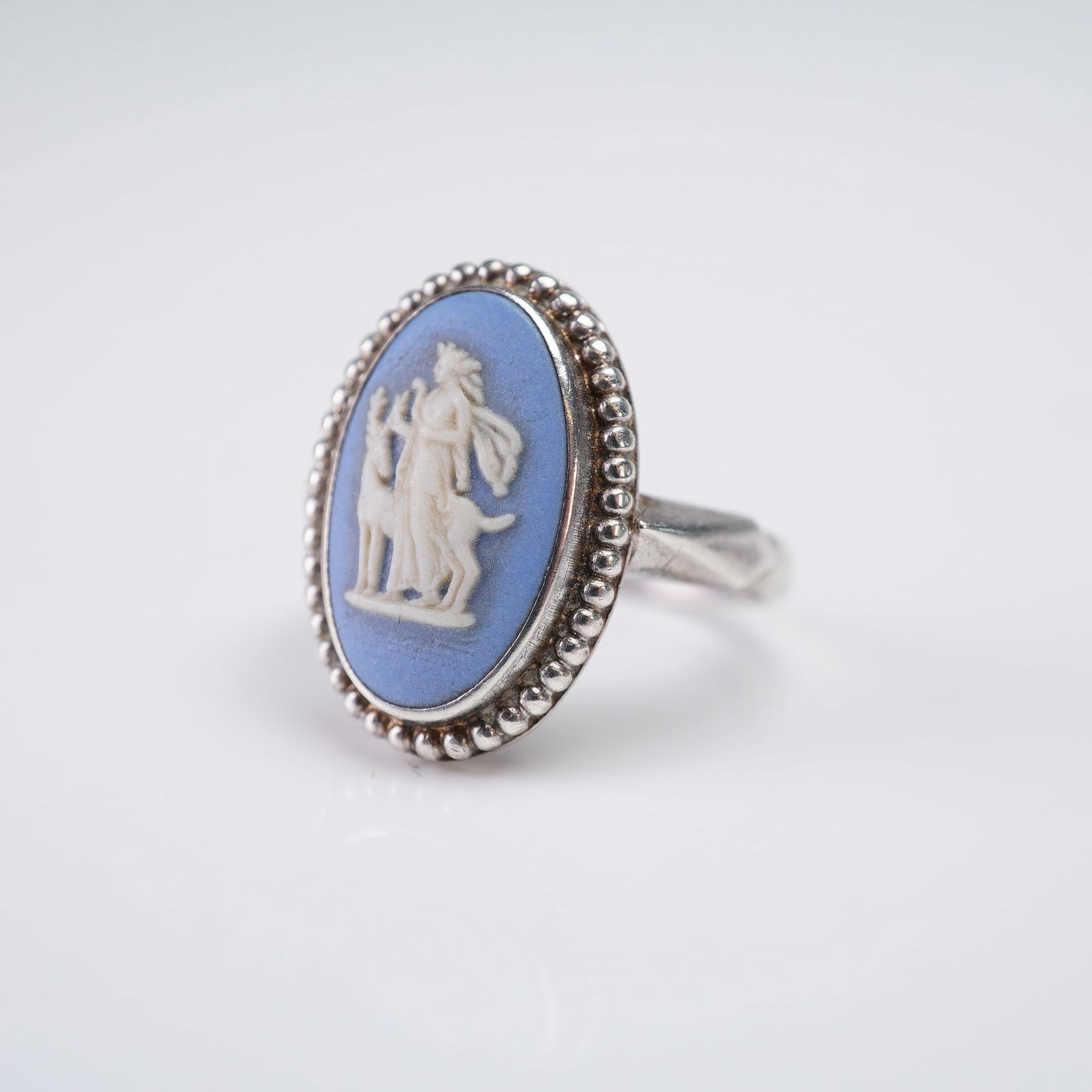 Wedgwood Blue Jasperware Cameo Sterling Silver Ring, Diana and the Stag: A sterling silver ring featuring an oval ceramic cameo insert. The cameo displays a white relief depiction of Diana the Huntress (Artemis) standing beside a stag, set against a pale blue ground. The s
