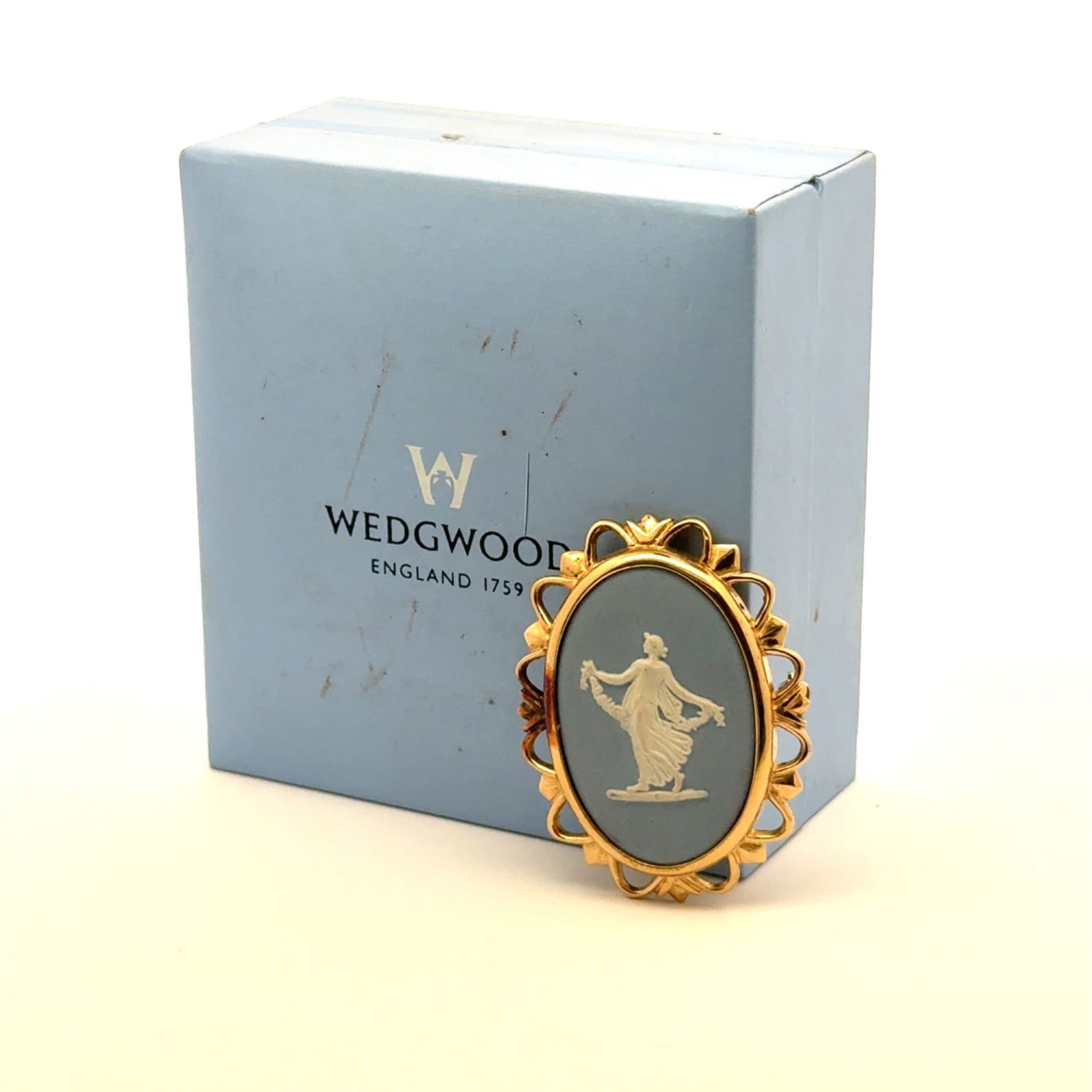 Wedgwood Blue Jasperware Cameo Gold-Tone Brooch, Dancing Hour: A Wedgwood Blue Jasperware cameo brooch and its original presentation box. The oval cameo features a light blue ground with a white Neoclassical relief figure depicting a dancing woman holding