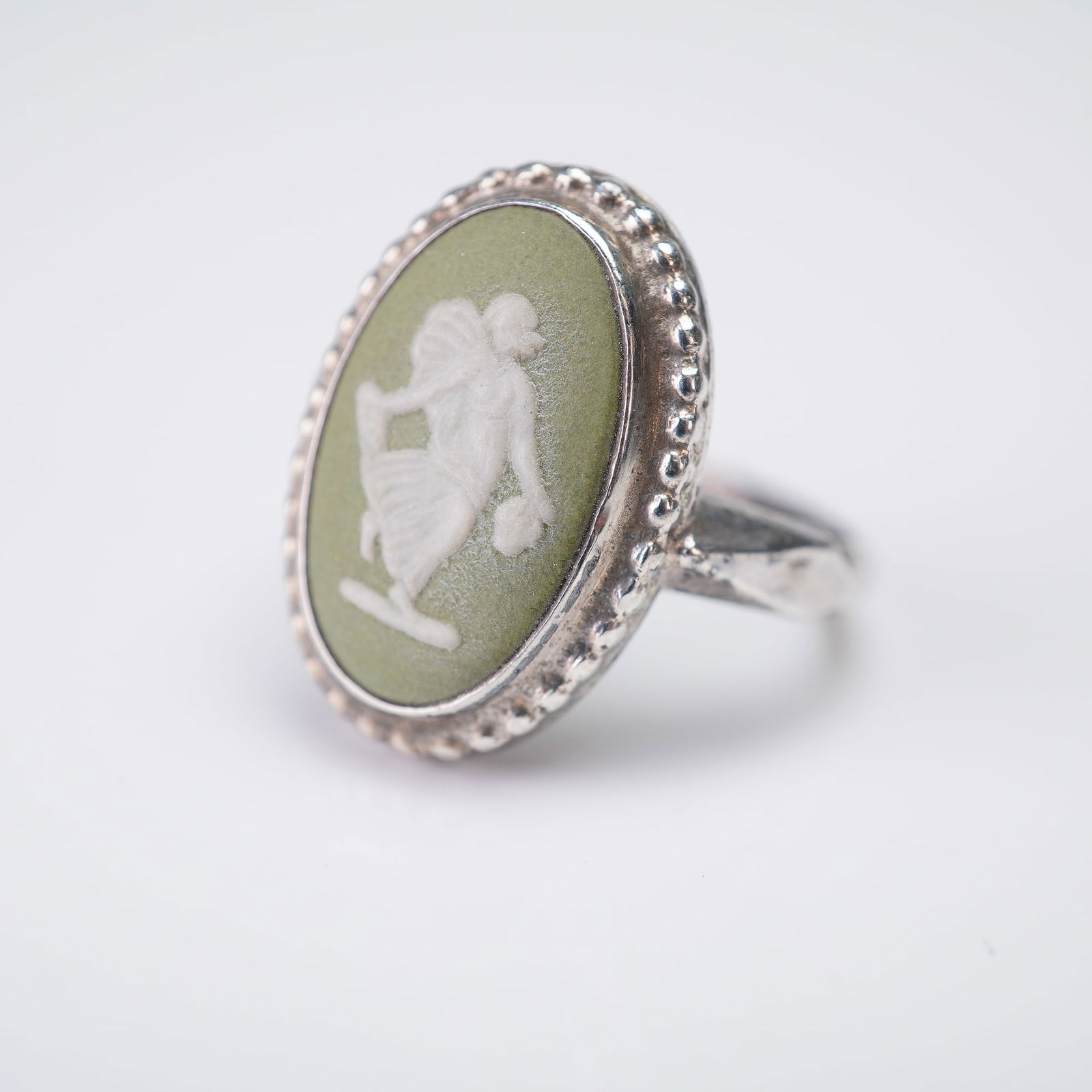 Wedgwood Green Jasperware Cameo Sterling Silver Ring: A ring featuring an oval cameo setting of Wedgwood Jasperware. The central element is a green ceramic plaque showcasing a white relief classical female figure dancing. The cameo is held in a sterling