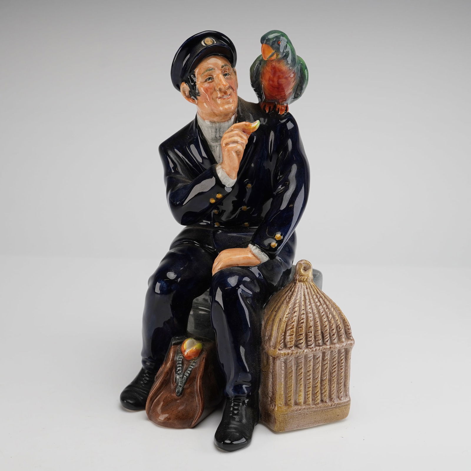Royal Doulton Shore Leave HN 2254 Porcelain Figurine: A single glazed porcelain figurine by Royal Doulton, titled "Shore Leave" and modeled as HN 2254. This figurine depicts a sailor in a deep navy blue uniform and cap, seated on a grey brick base, feedi