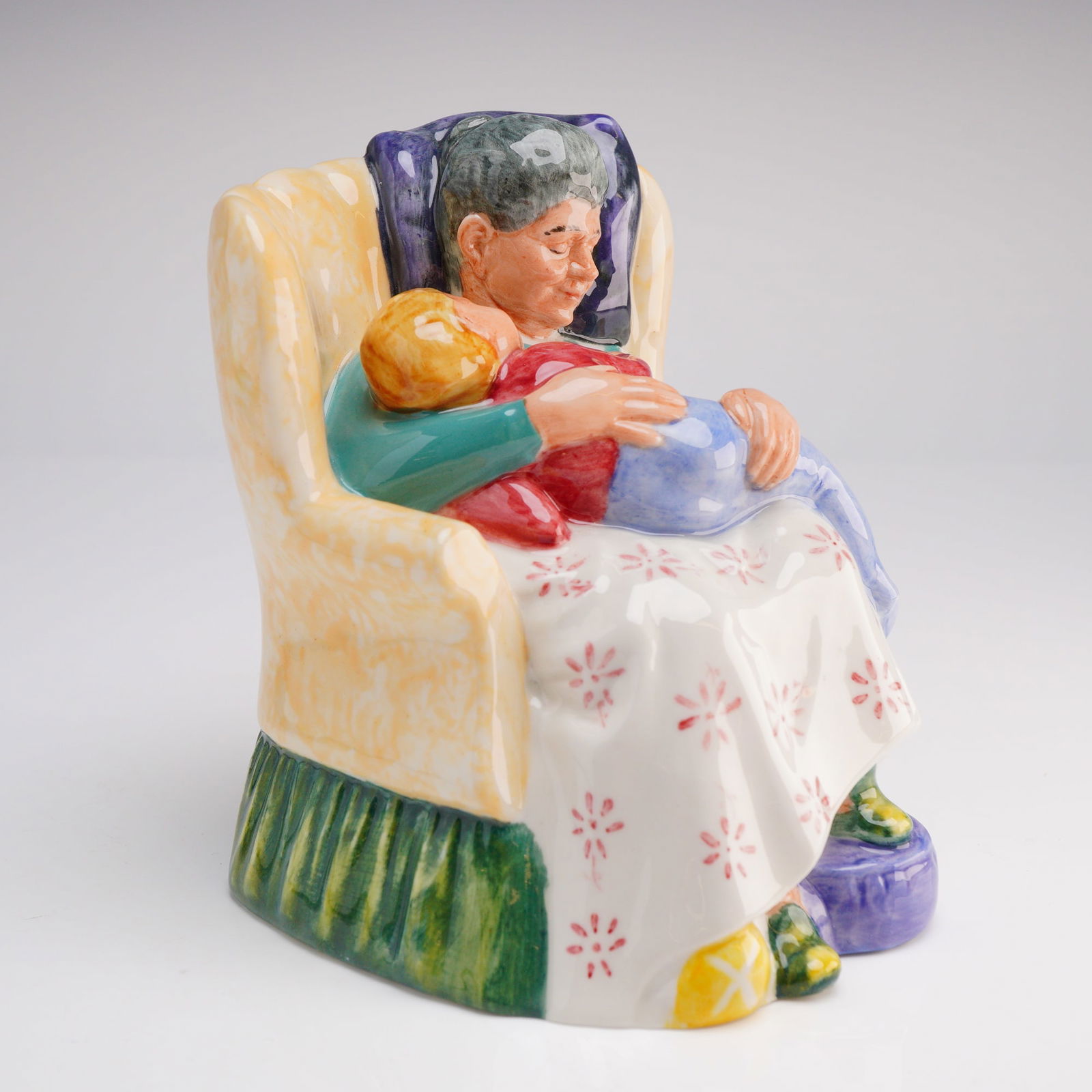 Royal Doulton Figurine Sweet Dreams HN2380: The piece depicts an older woman seated in a high-backed, butter-yellow armchair, cradling a sleeping child in her lap. The figurine is rendered in vibrant polychrome enamels, featuring a white skirt