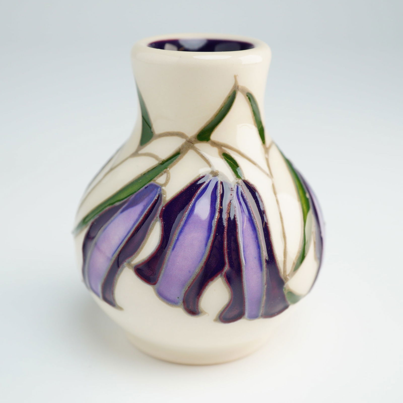 Moorcroft Pottery Miniature Vase, Bluebell: The vessel is decorated using the tube-lining technique, depicting purple bell-shaped flowers and green foliage against a cream ground. The interior of the vase is finished with a deep purple glaze. T