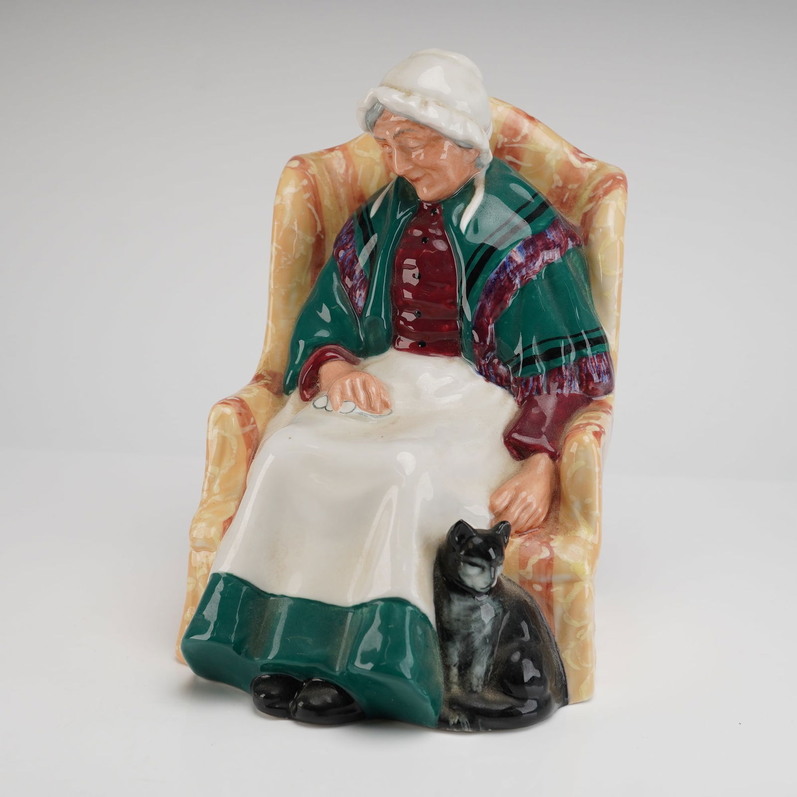 Royal Doulton Figurine Forty Winks HN 1974: The piece is executed in glazed porcelain or bone china and depicts a character study of an elderly woman seated in a patterned yellow and orange upholstered armchair. She wears a white bonnet, a deep