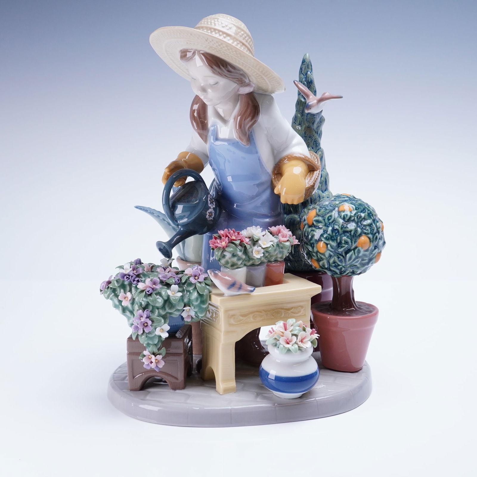 Lladro In My Garden Figurine 01008663, Signed: The figurine depicts a young female gardener, dressed in a white blouse, blue apron, and straw hat, tending to an array of potted flora, including a large flowering bush, a small topiary, and an orang