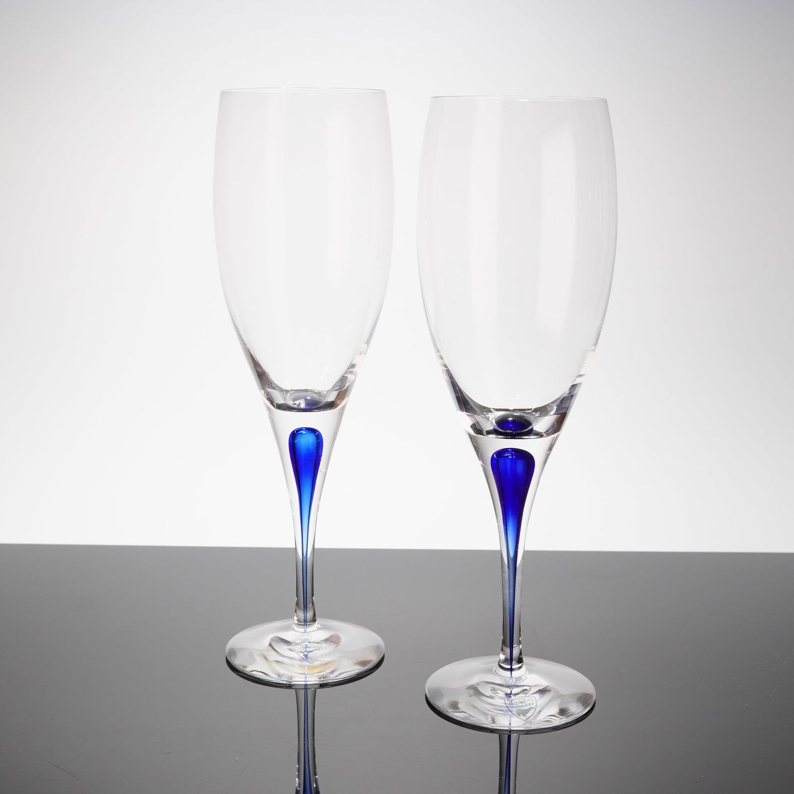 A Pair of Orrefors Intermezzo Blue Crystal Wine Glasses: A pair of stemmed glasses in the Intermezzo Blue patern, manufactured by Orrefors, Sweden. These items are crafted from clear crystal, featuring a slightly conical bowl supported by a solid stem. The