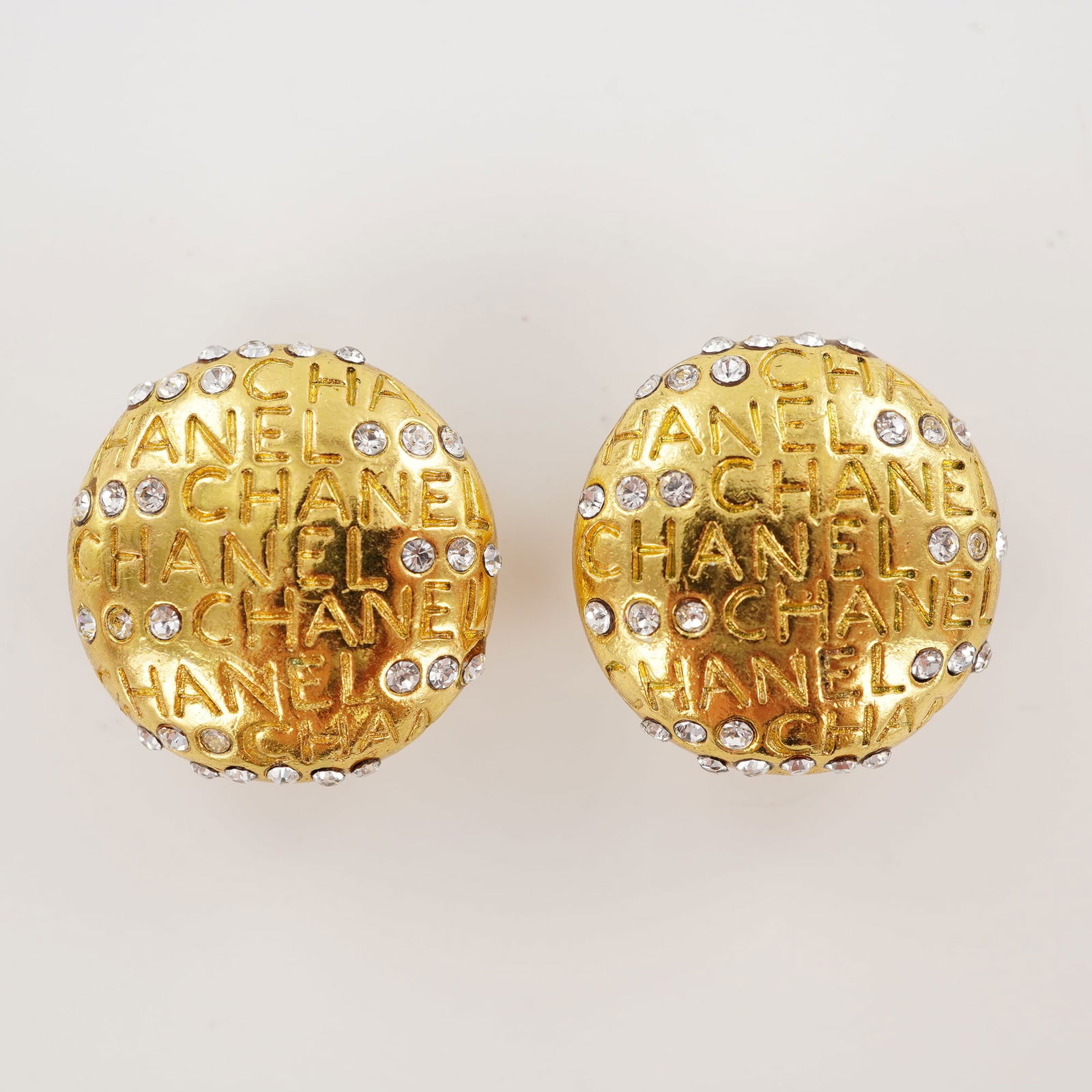 Chanel Logo and Rhinestone Gold Tone Clip Earrings: Designed in a domed profile, these Chanel earrings feature repeating CHANEL lettering across the surface with inset clear rhinestones. The earrings are made in gold tone metal and fitted with clip bac