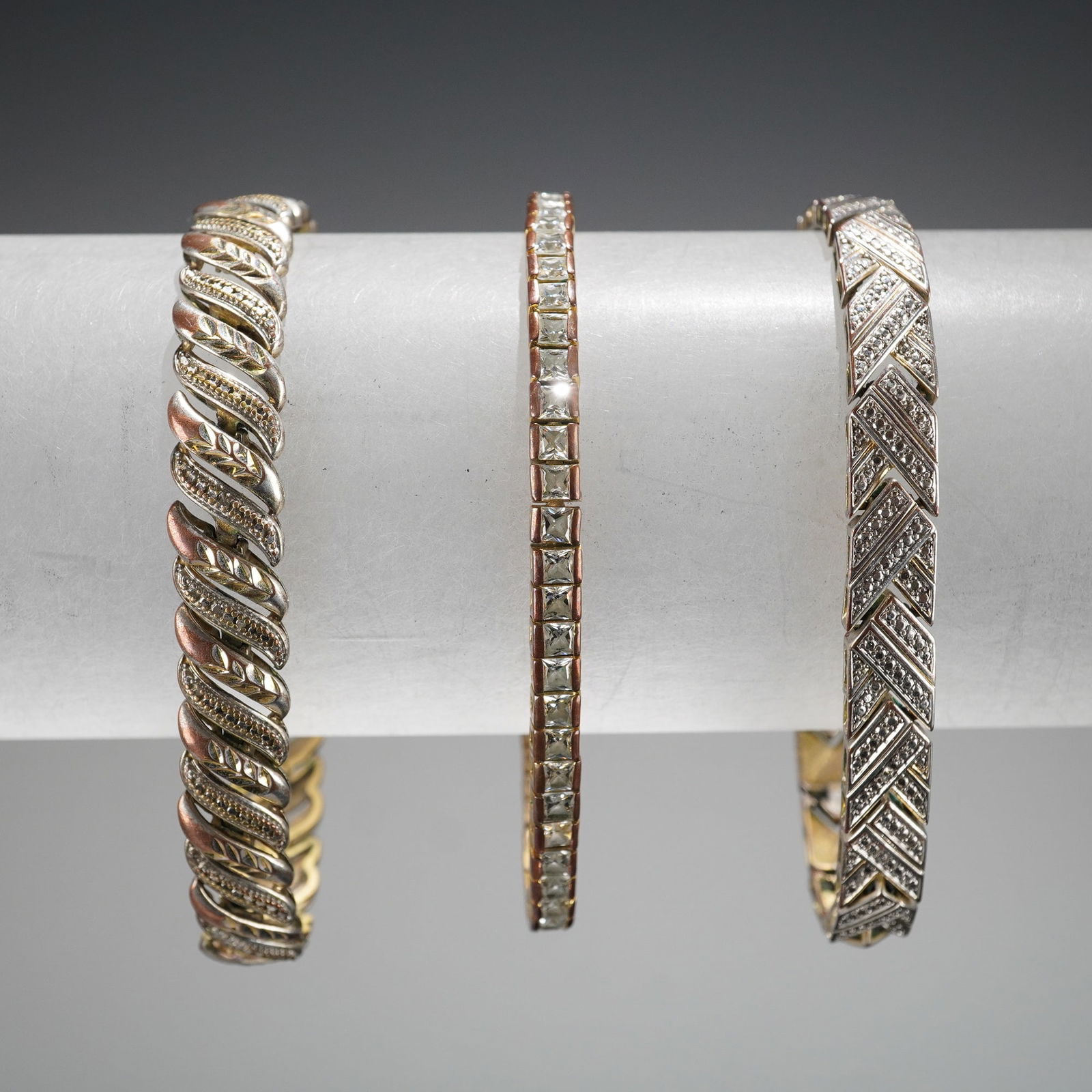 Trio of Contemporary Silver-Tone and Multi-Tone Costume Bracelets: A trio of contemporary costume bracelets constructed from plated base metal and set with colorless simulated stones. The collection includes three distinct styles: a wide, intertwined braided bracelet