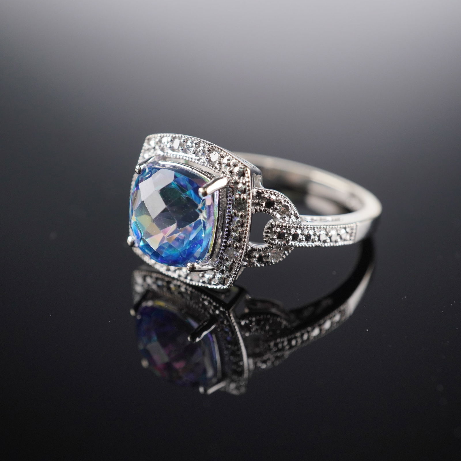 10K White Gold Blue Mystic Topaz and Diamond Ring: Set with a cushion-cut blue mystic topaz at the center, accented by twelve round brilliant-cut diamonds, eight framing the topaz and two on each shoulder. Crafted in 10K white gold and marked 10K HDS