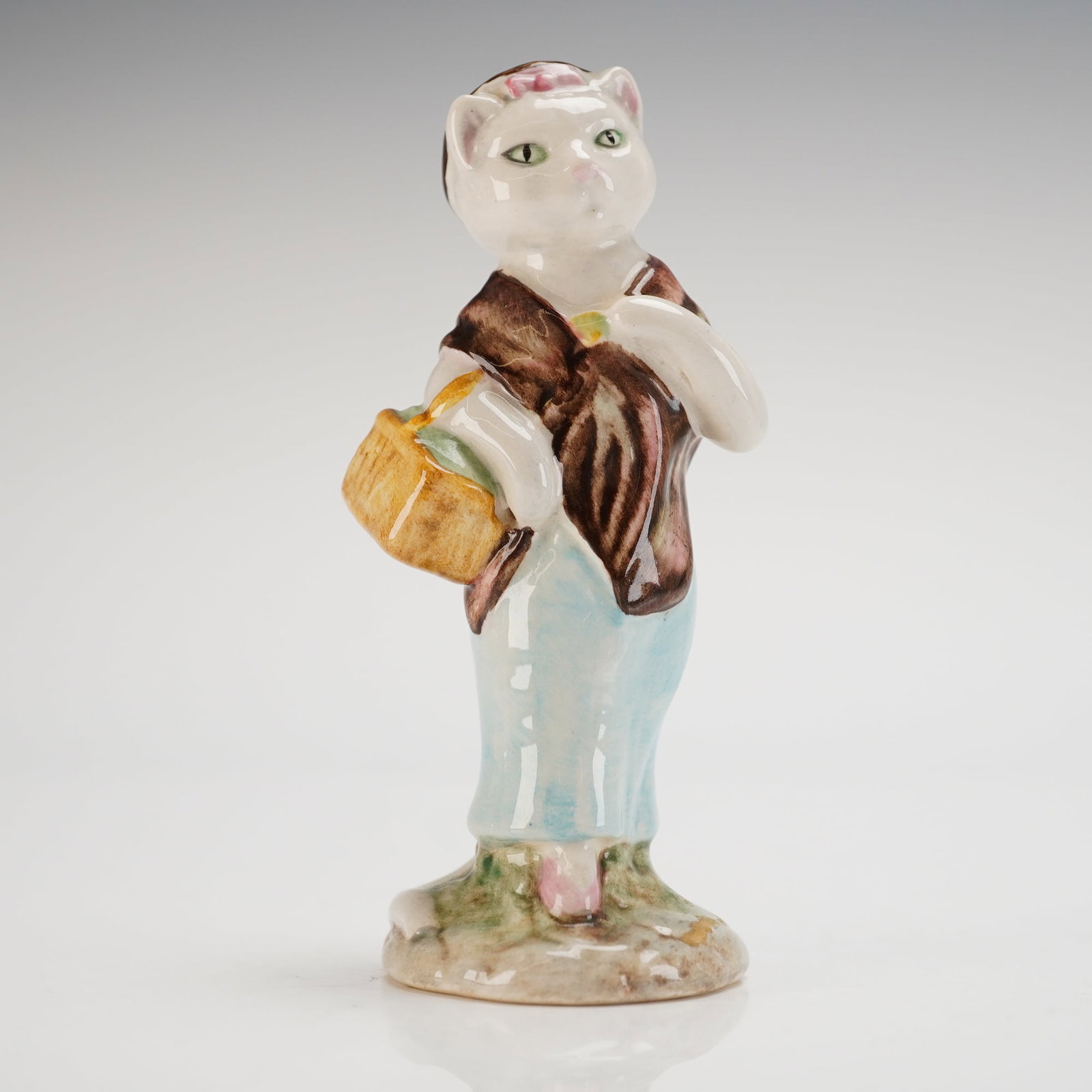 Beswick Beatrix Potter's Figurine, Susan: Model number P2716 portrays an elegant cat dressed in a pale-blue gown with a brown shawl and a cap accented by a pink bow. She holds a basket and stands atop a gray-green cobble-style base. The under