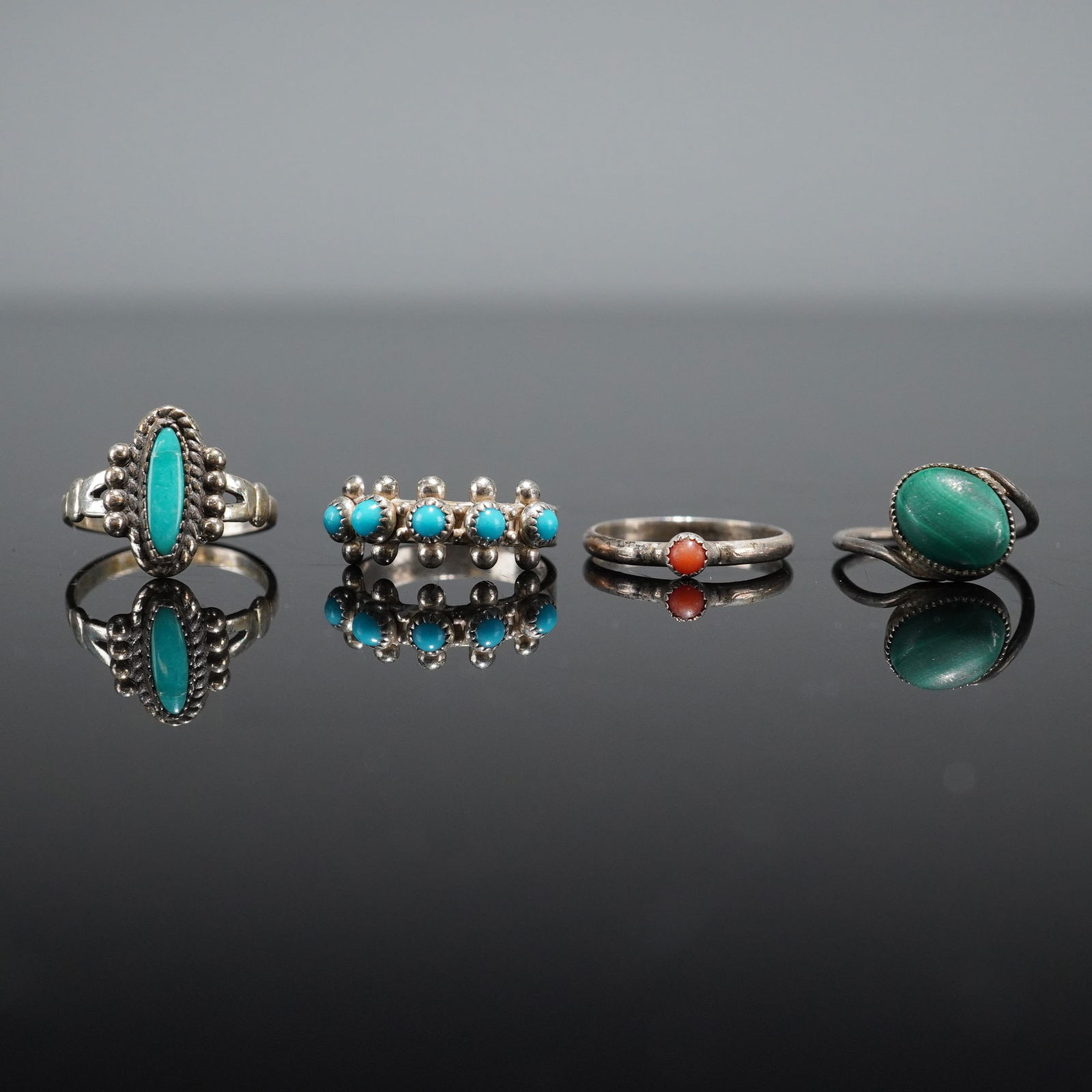 4pc Sterling Silver Rings with Turquoise Coral and Malachite: A collection of four sterling silver rings executed in a Southwestern style that includes four distinct designs, each set with various stones. The first ring features a large, elongated oval