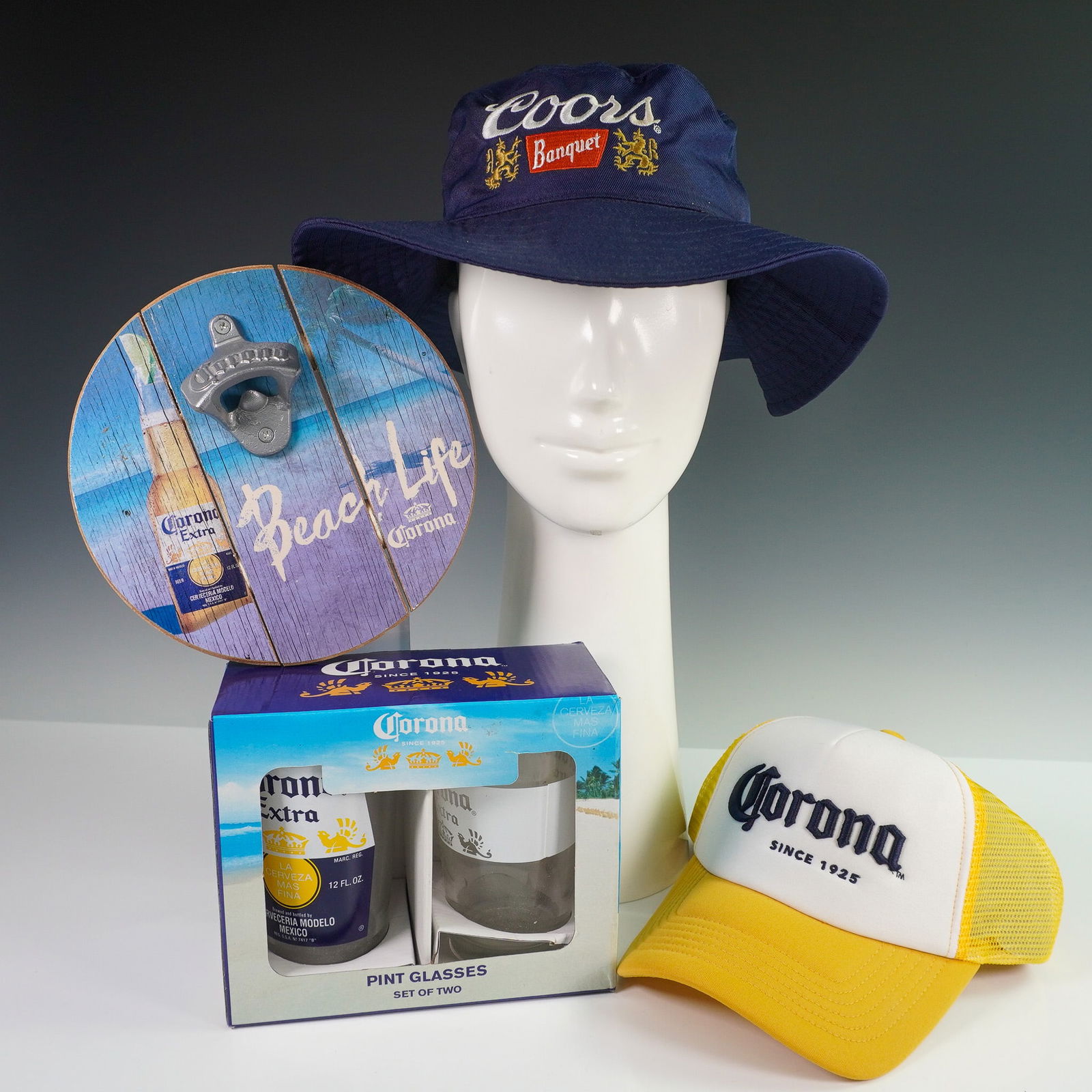 4pc Corona and Coors Hat Set and Barware Set: This 4 piece collection includes a mix of branded barware and apparel, featuring a round wooden Corona "Beach Life" wall mounted bottle opener, a boxed set of two Corona Extra pint glasses, a yellow a