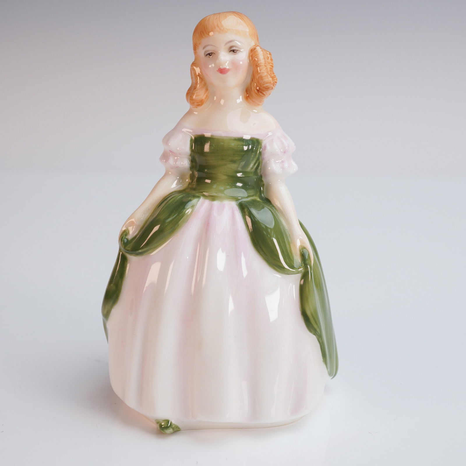 Royal Doulton Figurine, Penny HN2338: This delightful bone china figurine from Royal Doulton, titled Penny (HN 2338), depicts a young girl in a classically styled green and pale pink dress with softly gathered details. Designed by artist