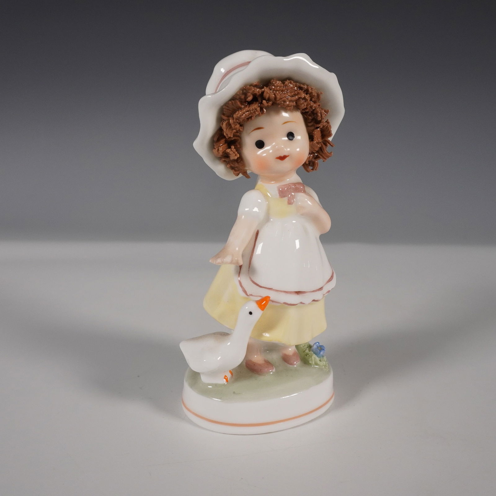 Napco Ware Bone China Figurine, Girl with Goose: A vintage Napco Ware bone china figurine featuring a young girl in a yellow dress with a white apron and a wide-brimmed hat, playfully interacting with a white goose. The figurine is finely detailed,