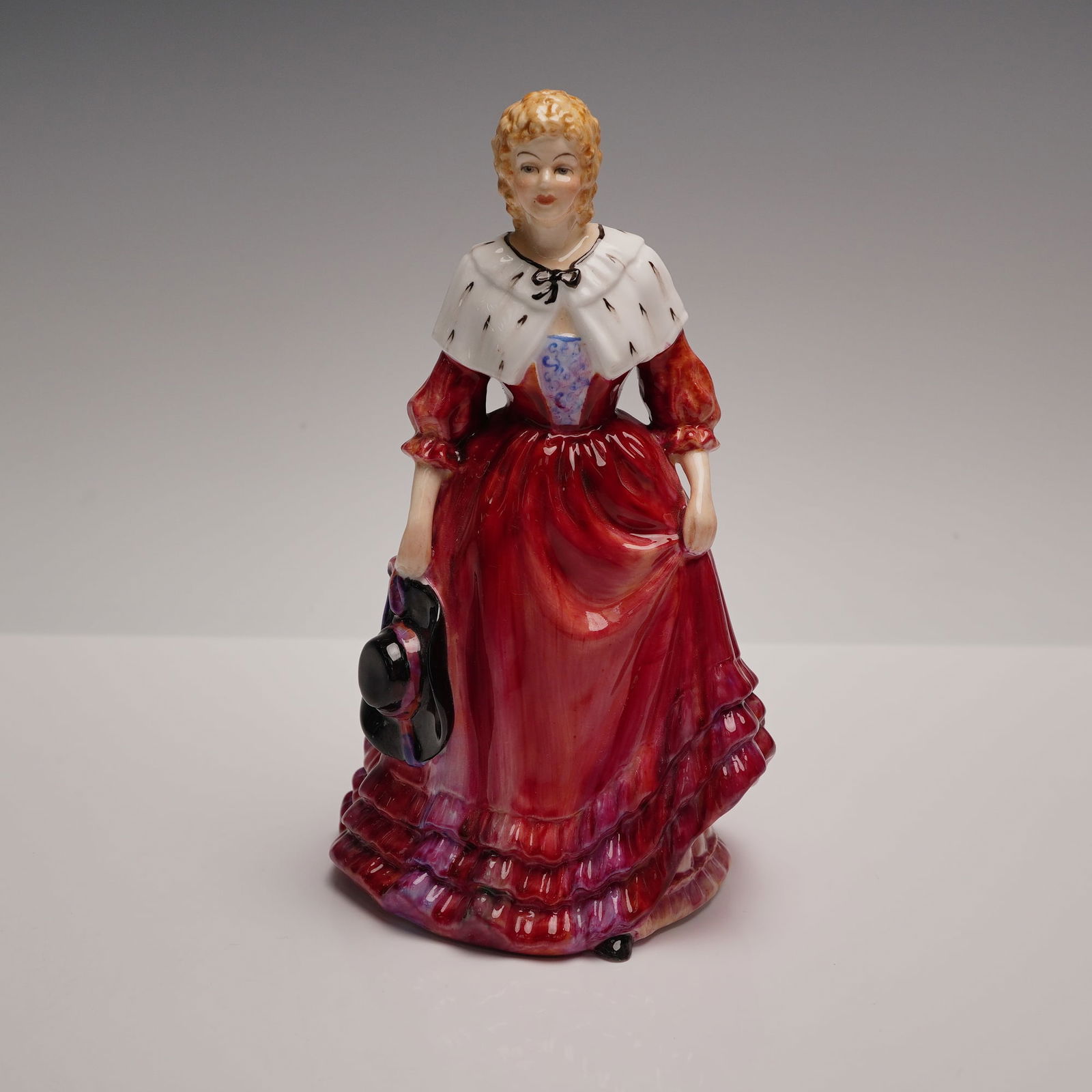 Paragon Bone China Figurine, Lady Cynthia: An elegant hand-painted bone china figurine titled Lady Cynthia, produced by Paragon, England. Modeled in rich crimson red and violet hues, this refined lady stands demurely holding her bonnet by her
