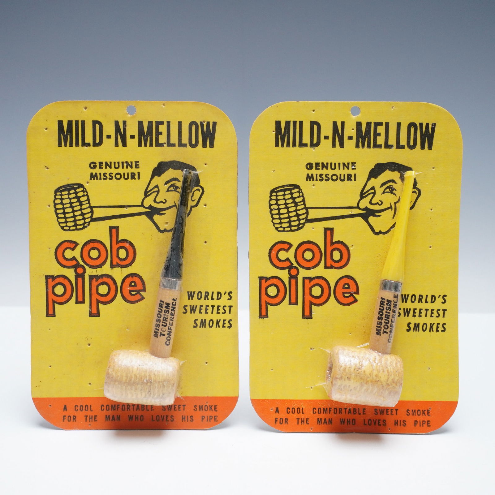 2pc Buescher's Mild-N-Mellow Missouri Cob Pipes: A pair of cob pipes each presented on its original yellow and orange display card featuring the classic slogan "Worlds Sweetest Smokes" and caricature branding. These genuine Missouri-made pipes refle