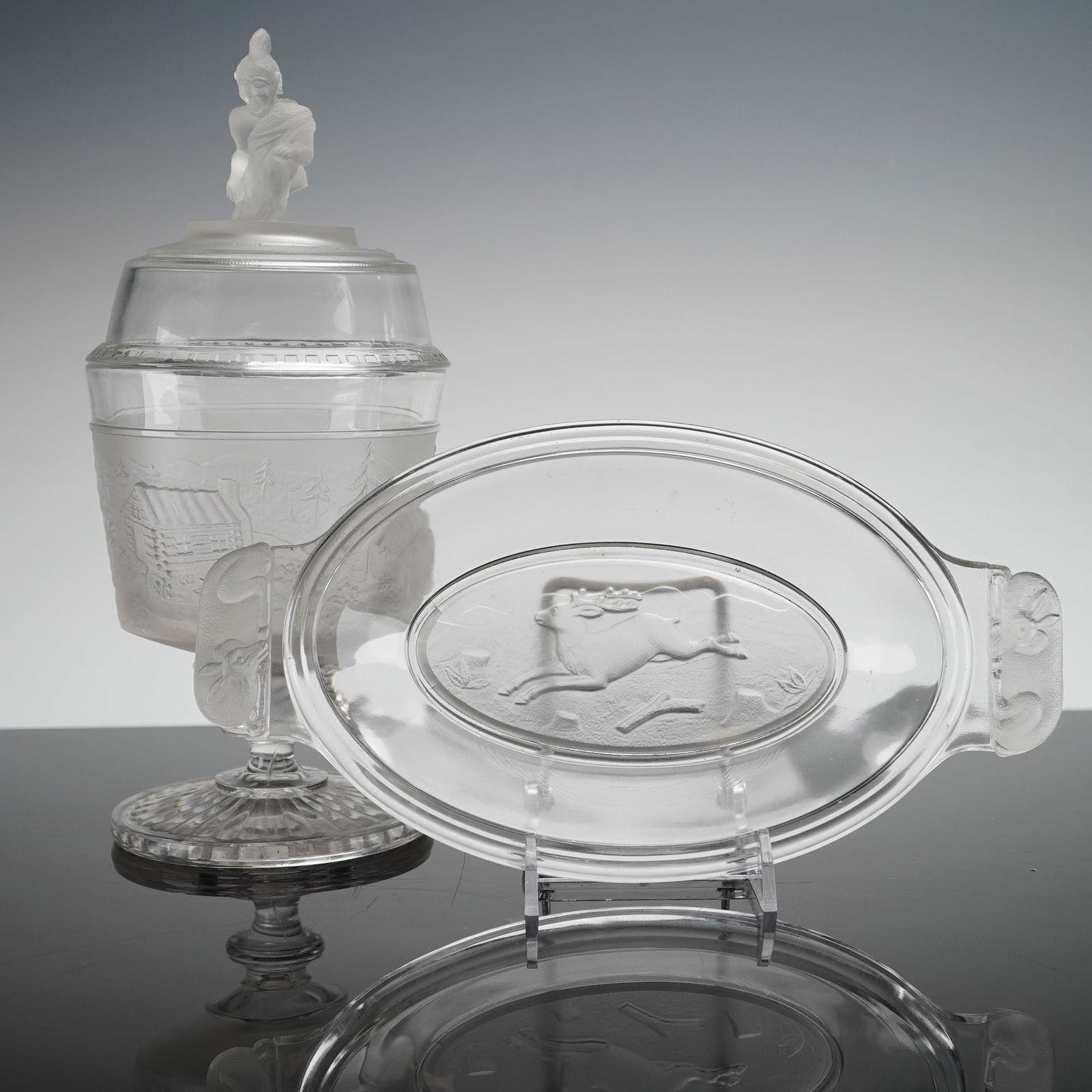 2pc Gillinder & Sons EAPG Pressed Glass Lidded Compote & Dish, Westward Ho: The grouping includes a covered compote with lid and an oval serving dish. Both items utilize clear pressed glass with extensive frosted relief molding. The covered compote stands on a ribbed pedestal