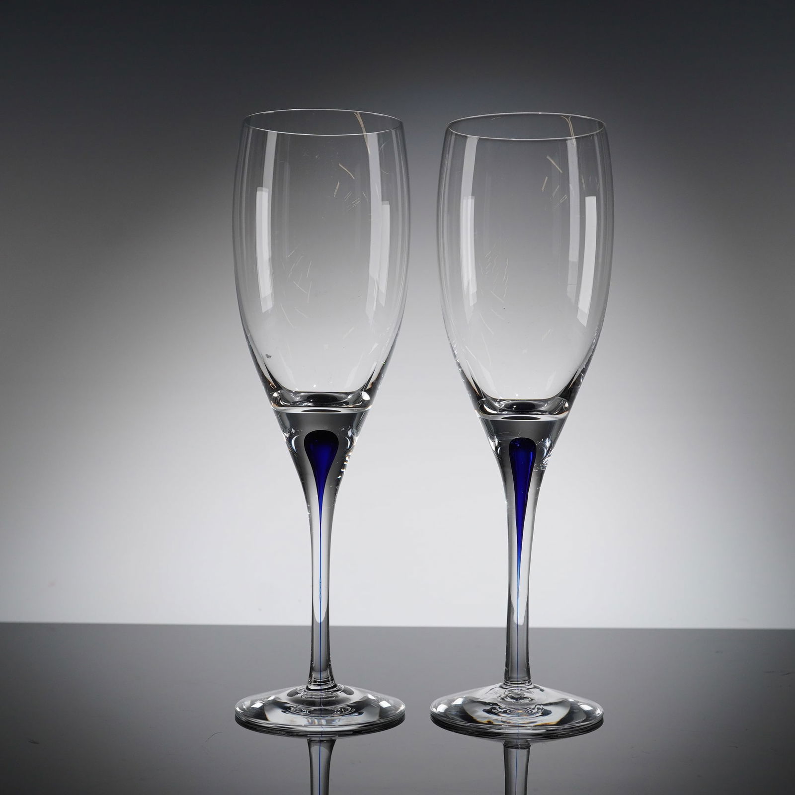 A Pair of Orrefors Intermezzo Blue Champagne Flutes: A pair of modern champagne flutes in the Orrefors Intermezzo Blue patern. Each fi?ute is constructed from clear glass with a slender stem and a tall, tapering bowl. The defining characteristic is the