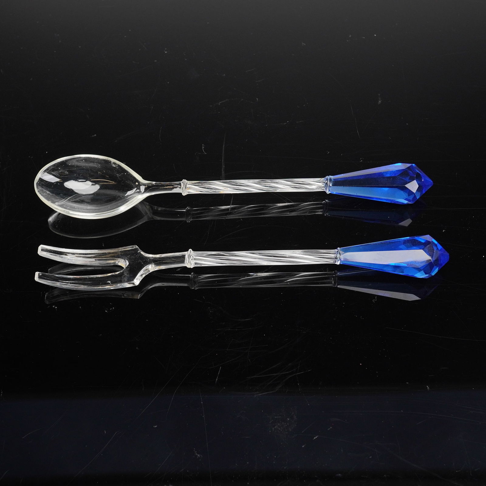 2pc Glass Serving Spoon and Fork with Cobalt Blue: Serving spoon and fork with cobalt blue tips, 20th-century decorative serving ware. Each utensil features a clear glass, spirally twisted stem leading to a large, faceted, drop-shaped finial crafted f