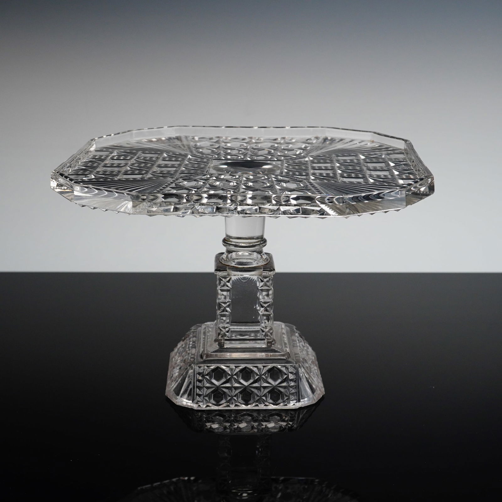 Adams & Co. EAPG Cake Stand, Valencia: The stand is crafted from clear glass and features an octagonal serving plate mounted on a short, multifaceted stem that rises from a tiered, squared base. The plate exhibits complex geometric cutting