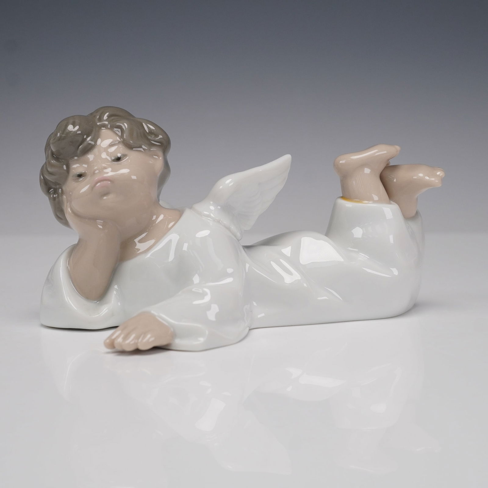 Lladro Porcelain Figurine, Angel Reclining 1004541: Glossy porcelain figurine by Lladro titled Angel Reclining, model number 1004541, sculpted by Fulgencio Garcia. Depicts a serene angel lying on its stomach, head resting in one hand and legs lifted be