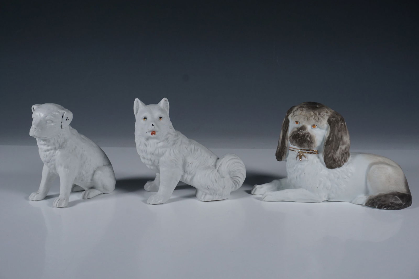 3pc Seated and Reclining Porcelain Dog Figurines: A set of three porcelain dog figurines, each uniquely crafted with fine detailing and hand-painted accents. The set includes a seated dog with black and gold highlights, a fluffy-tailed canine with su