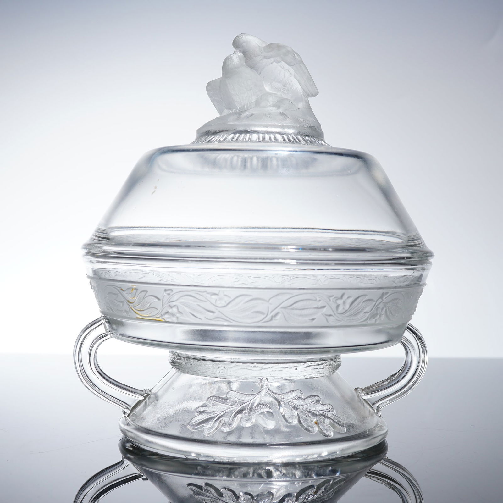 Gillinder Westward Ho Covered Compote: The piece is constructed of clear pressed glass and features a footed bowl with two applied handles and a matching dome-shaped lid. The bowl's body is decorated with a continuous band of stylized