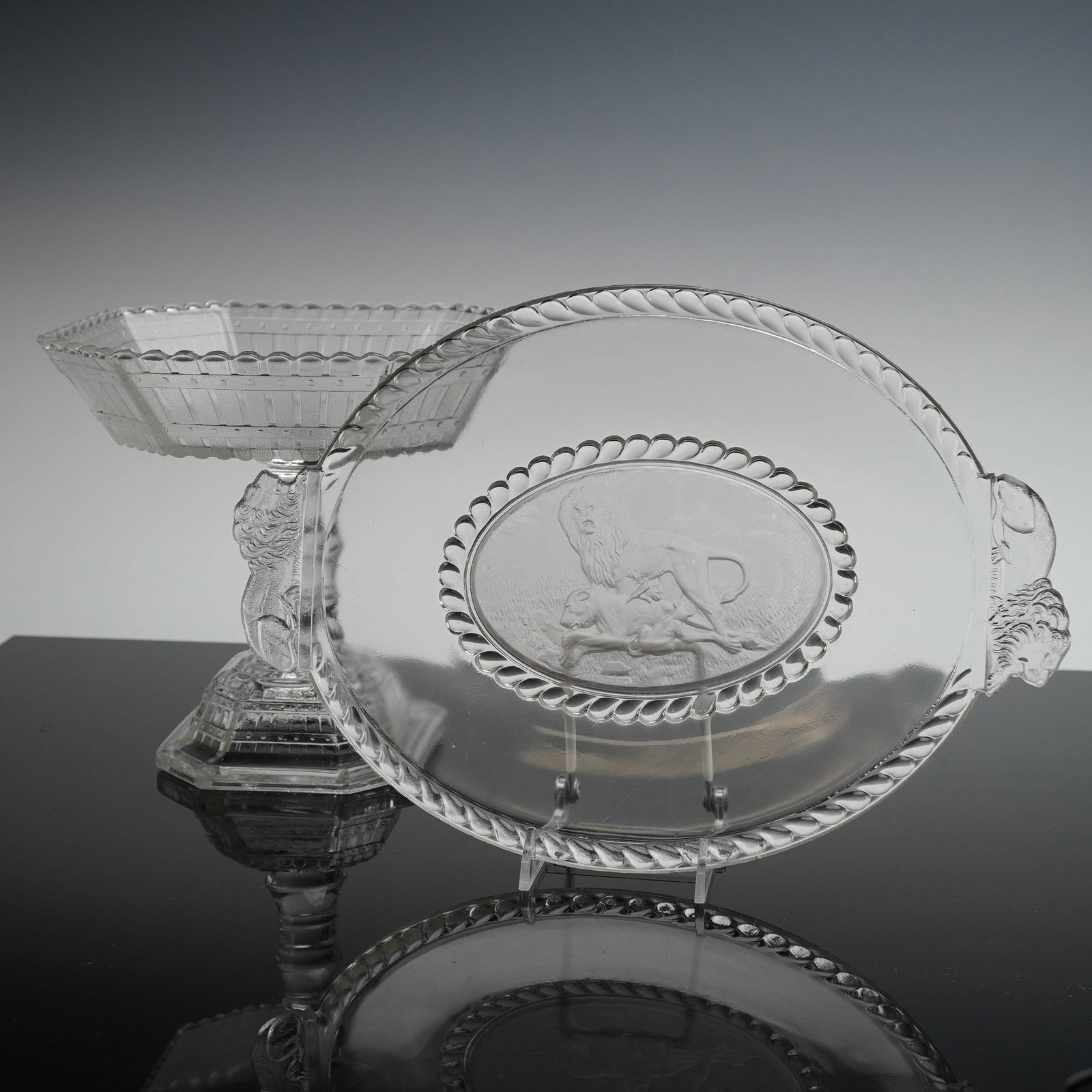 2pc Gillinder & Sons EAPG Oval Platter and Compote, Lion & Picket Fence: Both pieces are executed in clear pressed glass. The first item is an oval serving platter or tray, in the lion pattern. It features stylized figural lion head handles and a wide rope-twist border. Th