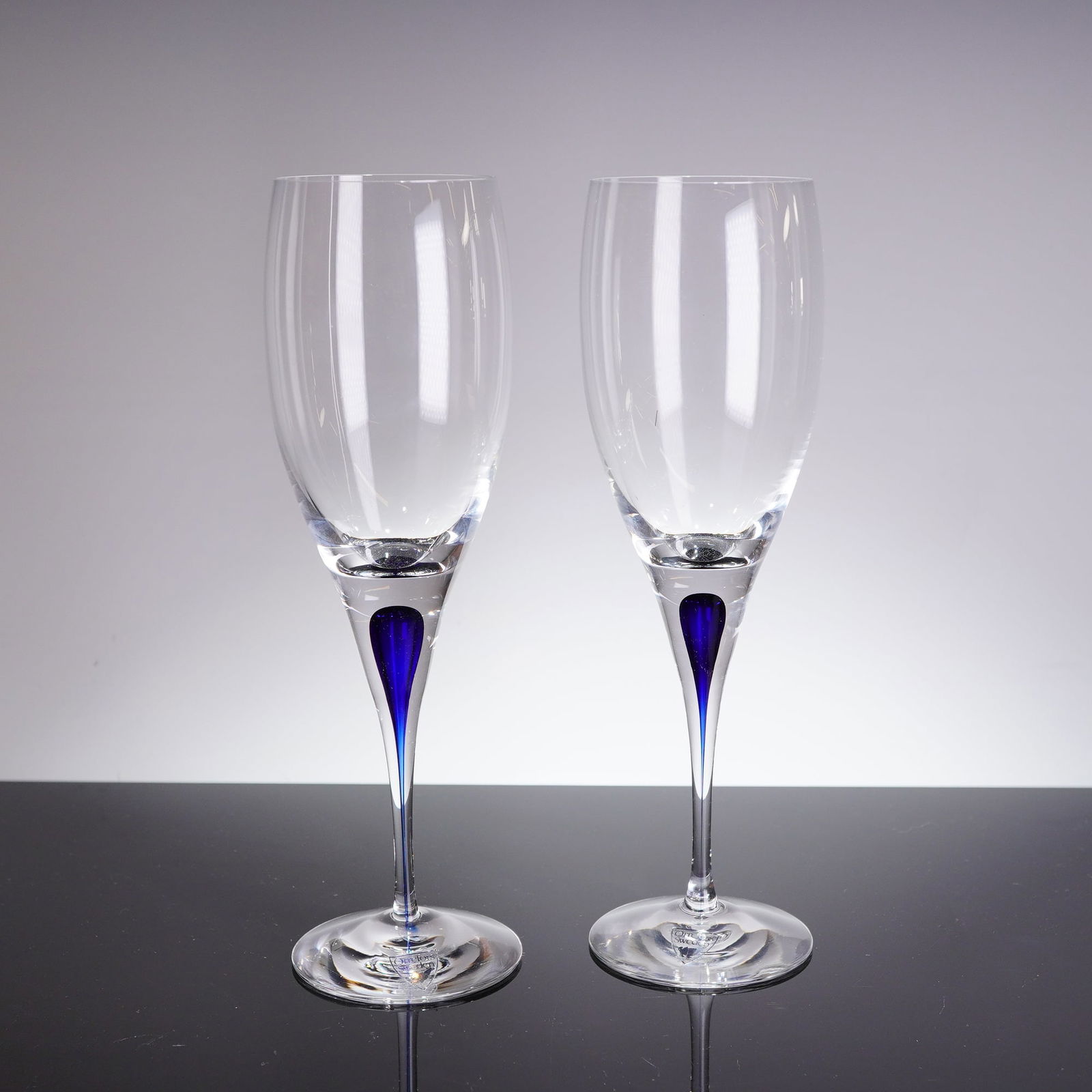 A Pair of Orrefors Intermezzo Blue Crystal White Wine Glasses: A Pair of Orrefors Intermezzo Blue crystal white wine glasses. These contemporary glasses are crafted from clear crystal, featuring a tall, slender bowl supported by a tapering stem and a clear circul