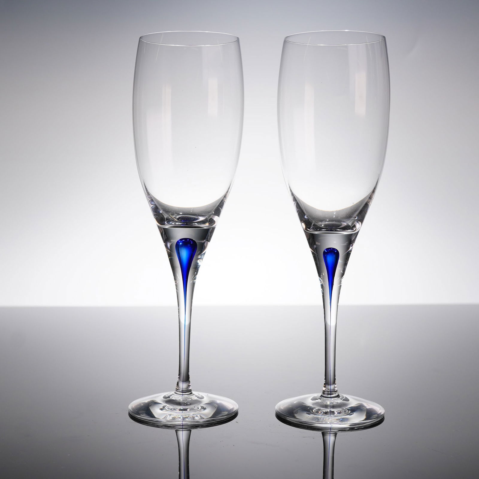A Pair of Orrefors Intermezzo Blue White Wine Flutes: A pair of atributed to Orrefors in the Intermezzo Blue patern. The stemware is fabricated from clear crystal, featuring a tall, narrow bowl suitable for sparkling wine, supported by a slender stem and