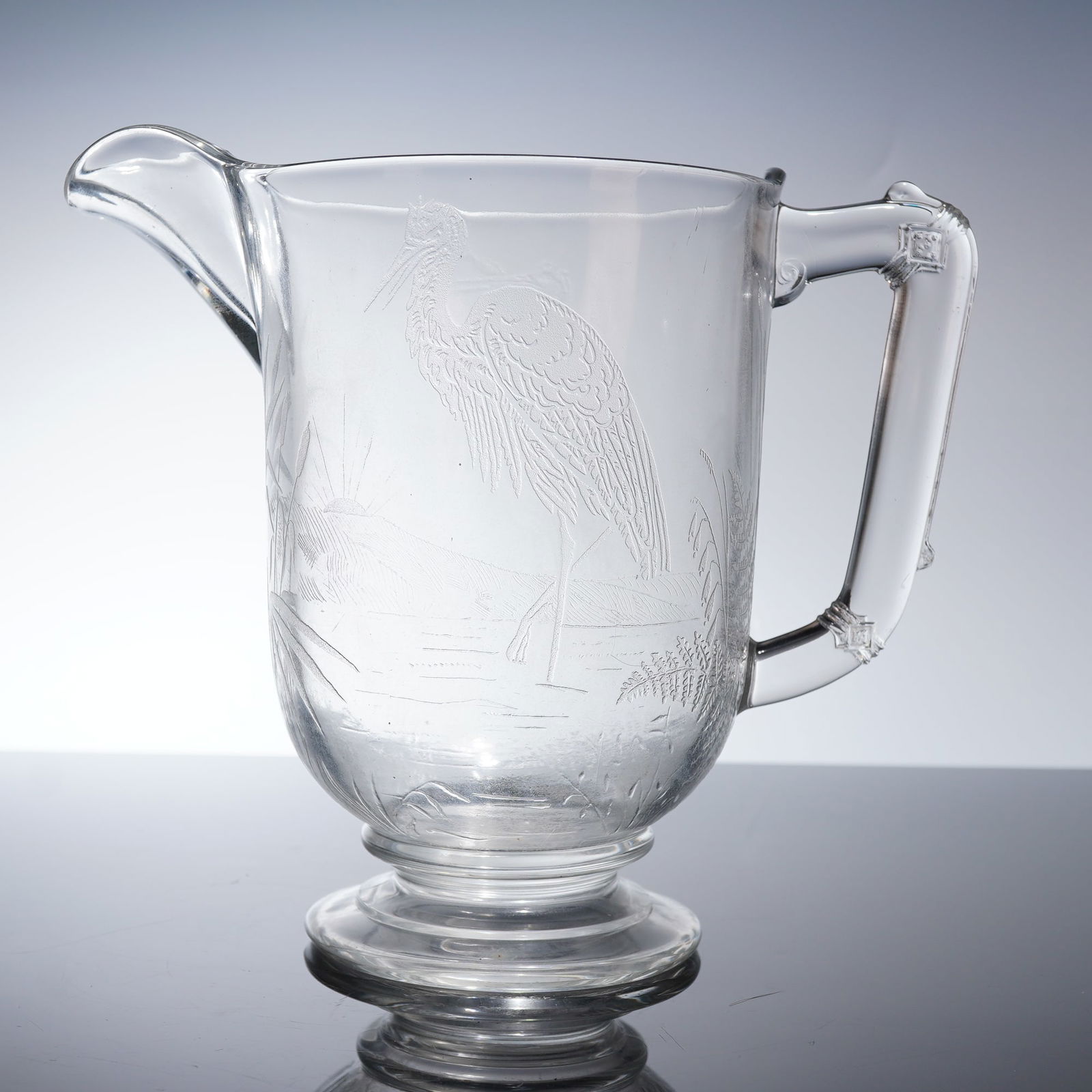 Dithering & Co. EAPG Pitcher, Wading Heron: The glass vessel features a slightly tapered body rising from a low, footed base. It has a pronounced pouring spout and an applied strap handle. The body is decorated with extensive acid-etched detail